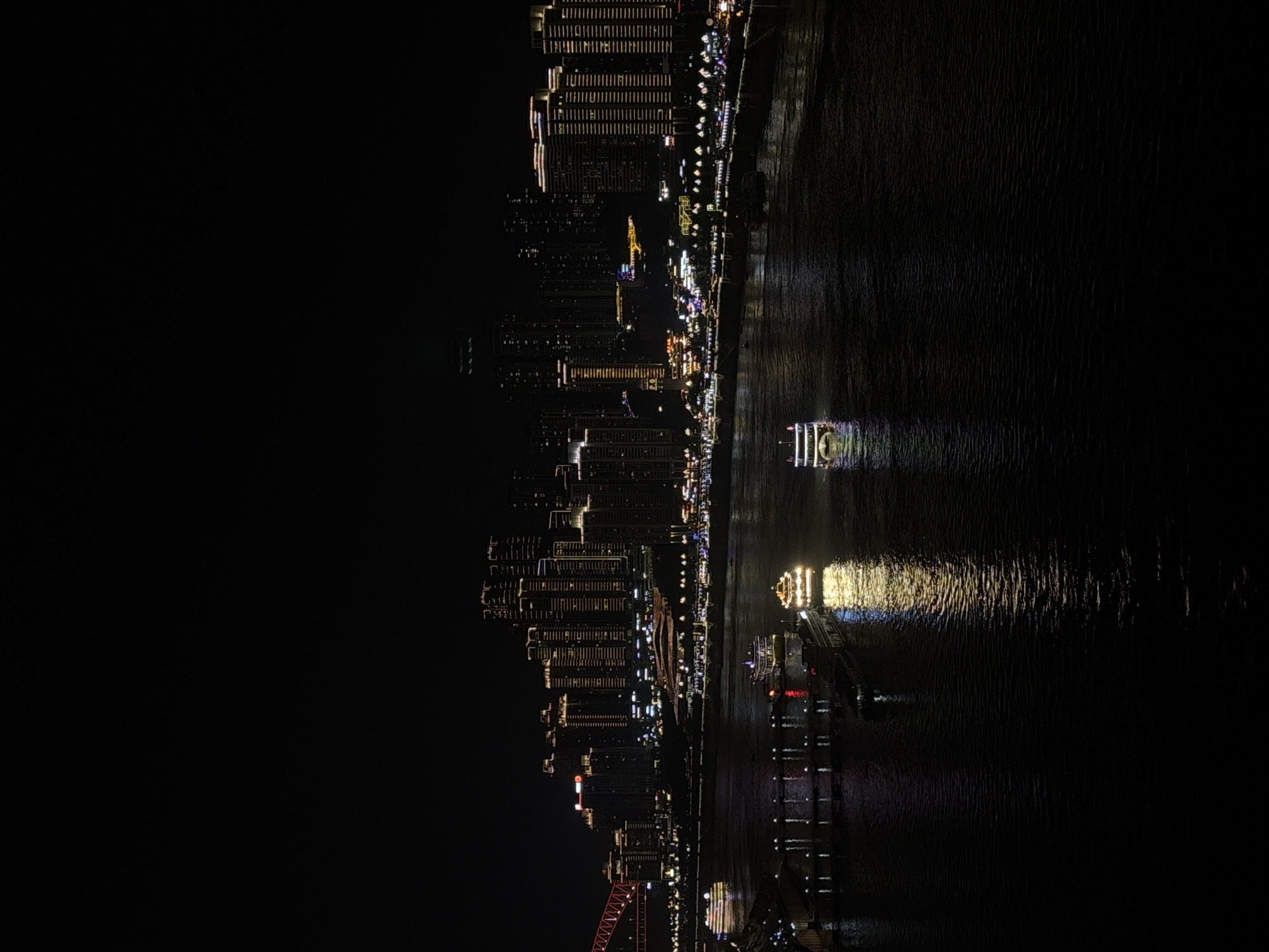 The image depicts a night view in Nan'an District, China, featuring a riverfront cityscape illuminated with lights. Skyscrapers and buildings line the riverbank, reflecting in the dark water. Two illuminated boats can be seen on the river, their lights creating streaks of reflection. The overall environment is dark, with the city lights providing the primary illumination. A red structure, possibly a bridge, is visible at the bottom of the frame.