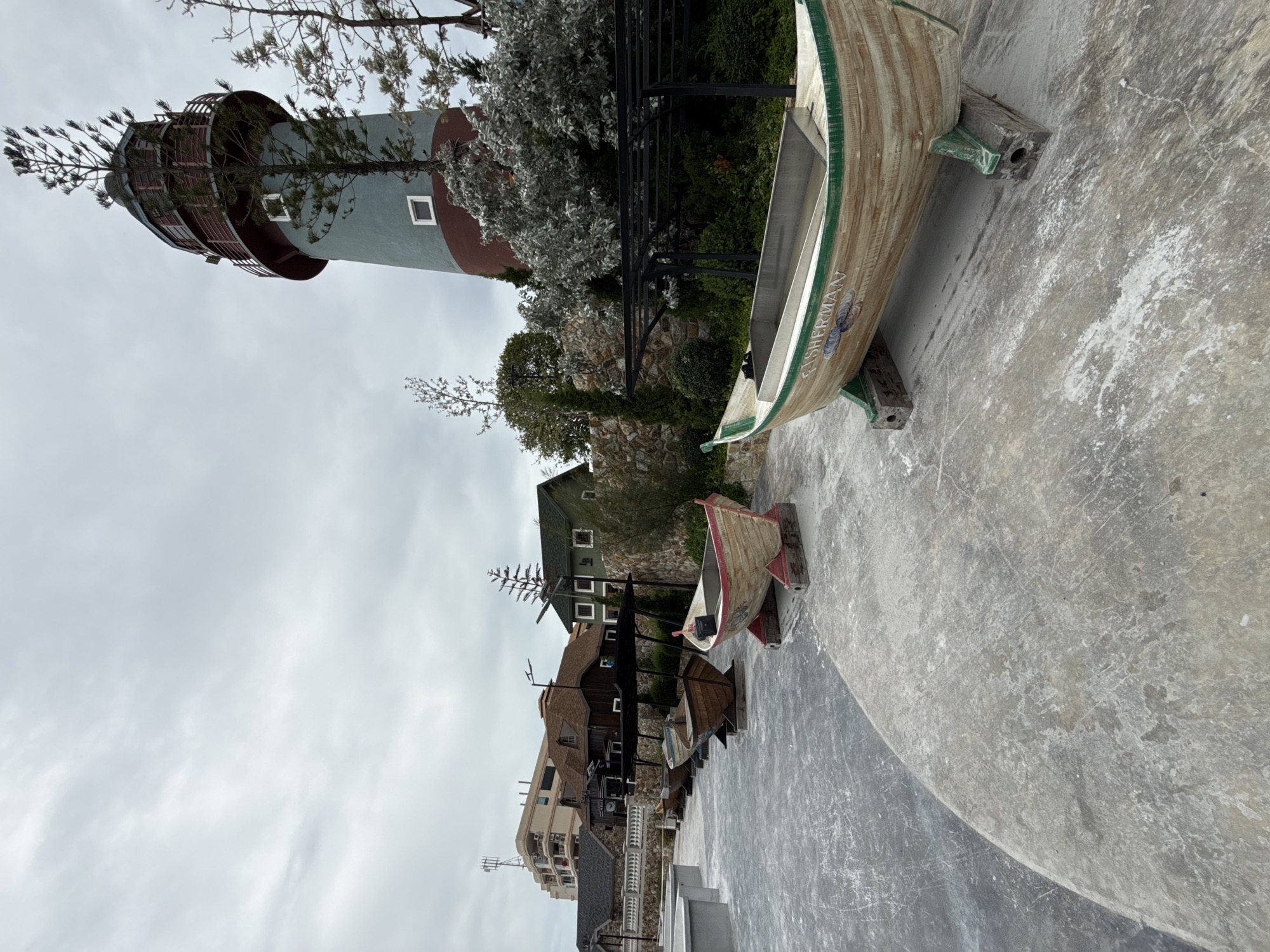 This image depicts an outdoor scene in Pattaya City, Thailand, featuring a lighthouse, several decorative boats, and a row of buildings in the background. The lighthouse, with its distinctive red and teal color scheme, stands tall against an overcast sky. In the foreground, four wooden boats are displayed on concrete stands. The closest boat is painted a light wood color with green trim and features the word "FISHERMAN" on its side. Behind it, three other boats are visible, each with a rustic appearance.
Beyond the boats, a cluster of buildings with pitched roofs and a contemporary multi-story building are situated. The architecture suggests a resort or a themed area. The ground is paved with concrete, showing some wear and tear. The environment is clean and well-maintained. There are no people visible in the image. The lighting is diffused due to the cloudy weather. The overall impression is one of a coastal-themed attraction or a picturesque tourist spot.