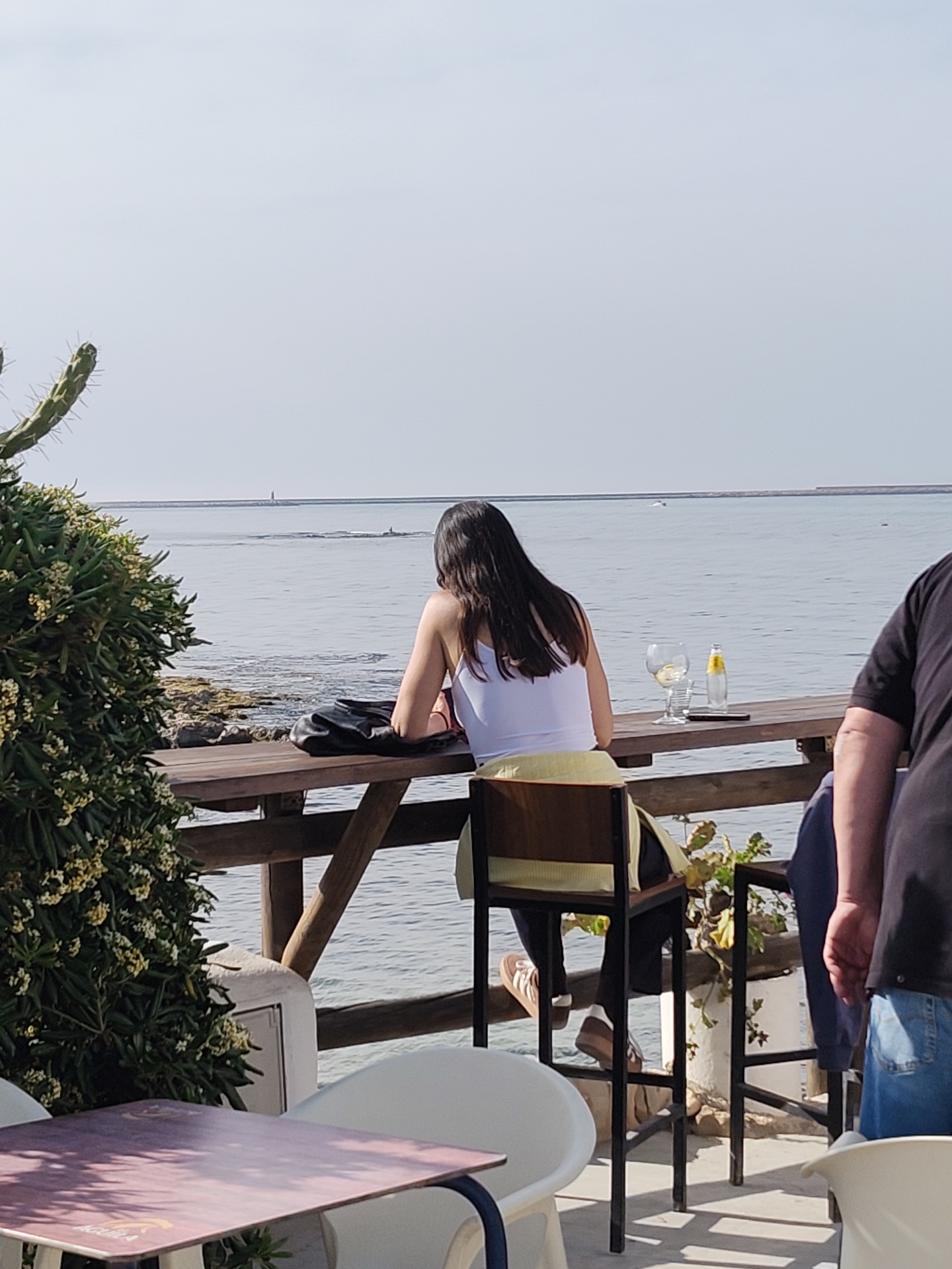 A medium shot captures a woman with long, dark hair sitting at a wooden table with a view of the sea in Dénia, Spain. She wears a white tank top and is positioned with her back to the viewer. On the table beside her are a glass containing a drink and a bottle. A black purse rests next to her. The table is part of a wooden railing structure overlooking the water.
To the left of the scene, a cactus-like bush with white flowers partially obscures the view. In the foreground, a table with a maroon and white top is surrounded by white plastic chairs. To the right, a man in a black t-shirt and jeans is partially visible. The sea stretches out in front of the scene towards the horizon under a clear sky. A breakwater or pier is visible in the distance.