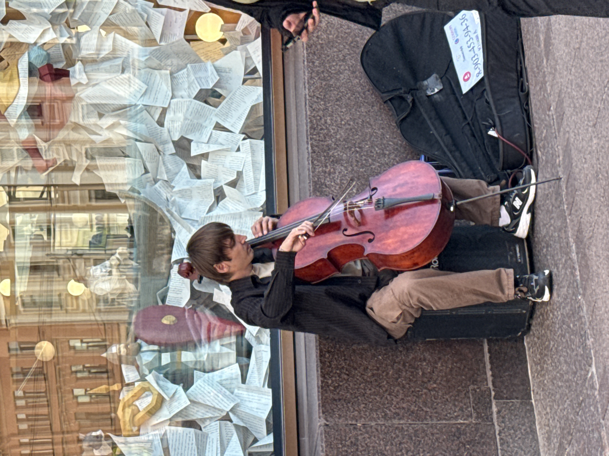 A male street musician is playing a cello in front of a shop window on Nevsky Prospekt in Saint Petersburg, Russia. The shop window is decorated with numerous sheets of paper, resembling old documents or sheet music, and some decorative elements. The musician is seated on a black amplifier, dressed in a dark shirt and brown trousers. His cello is a reddish-brown color. To his right, a black cello case is open on the ground, with a white sticker displaying a phone number and some text. Another person, partially visible, stands to the right, holding a mobile phone. The ground is paved with light-colored cobblestones.