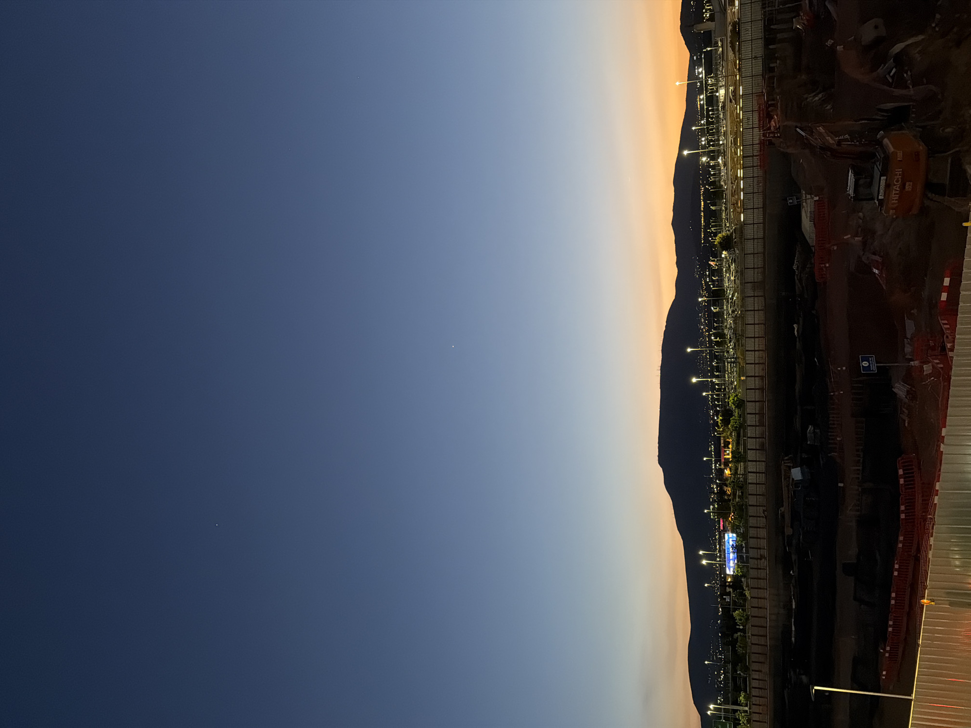 The image shows a view at dusk in Spata-Loutsa Municipal Unit, Greece. The sky is a gradient from dark blue to orange. The outline of a mountain range is visible. The foreground contains a construction site, including a Hitachi excavator, orange safety barriers, and a disabled parking sign. There are also streetlights visible.