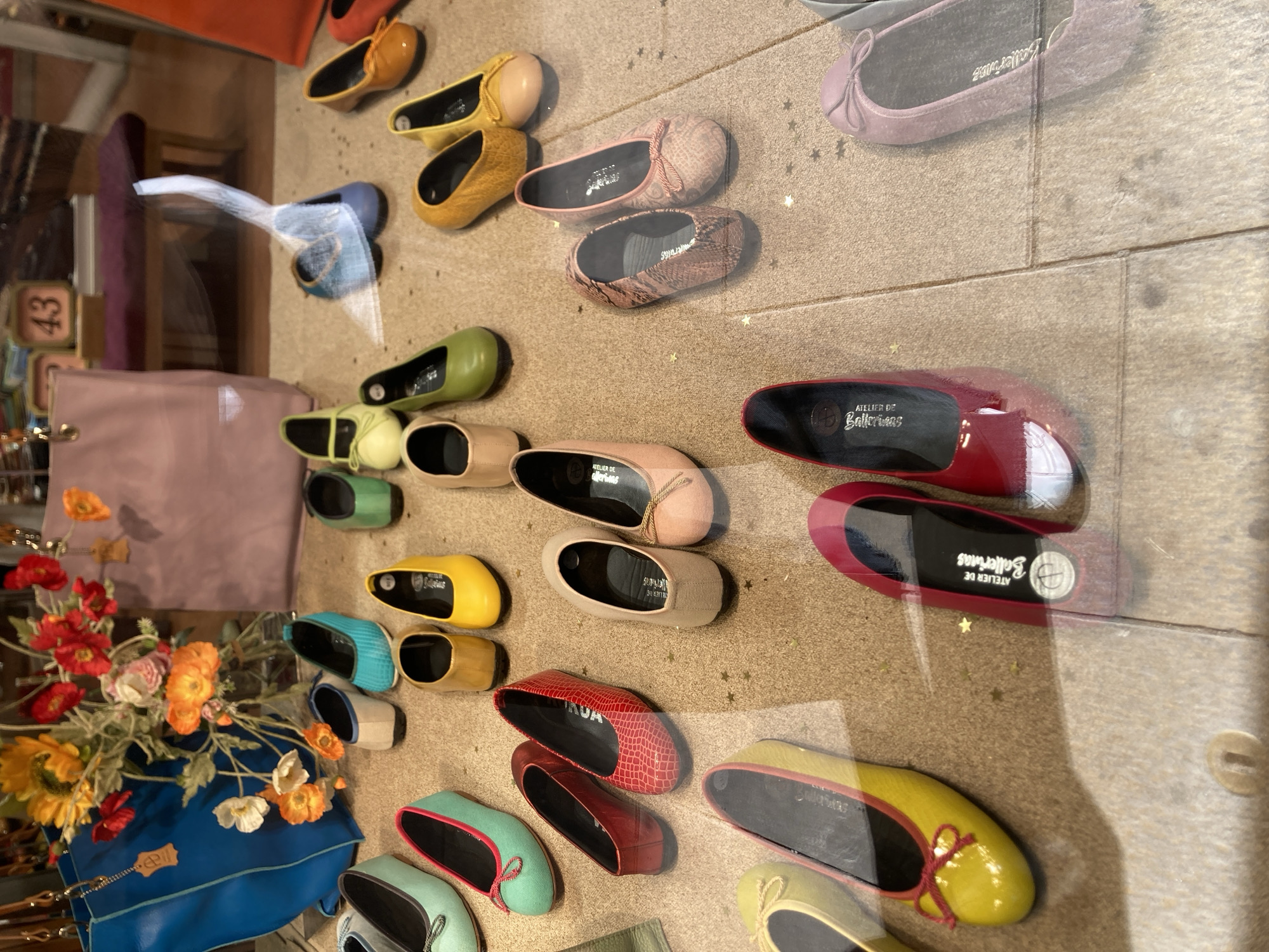 The image is a medium-angle, eye-level shot of a shoe store display window, likely located in Barcelona, Spain, given the context provided. The window showcases a variety of women's flat shoes in assorted colors and styles, arranged on a textured beige floor. In the background, a portion of a retail interior is visible, including a display of flowers, a pink handbag, and a bench with a dark cushion.
Dominating the foreground is a collection of flat shoes. Starting from the bottom left, a bright yellow flat shoe with a red bow is visible. To its right, a red flat shoe with a textured pattern and a matching red trim is displayed. Further to the right, a shiny red flat shoe with a white accent on the toe is present. Moving up, a pair of light pink flats with small bows are positioned centrally. To their left, a pair of beige flats with small bows are seen. Above these, there are several pairs of shoes in shades of green, yellow, brown, and red, as well as some light blue and turquoise flats.
Behind the shoes, a large, bright blue tote bag hangs to the left, adorned with a leather tag. A bouquet of flowers, featuring sunflowers, red poppies, and orange and pink blossoms, is arranged behind the blue bag. A pale pink handbag is partially visible in the upper right background. The overall impression is that of a neatly organized retail display aiming to attract customers. There are no people visible in the image. The lighting appears to be consistent with indoor retail lighting.