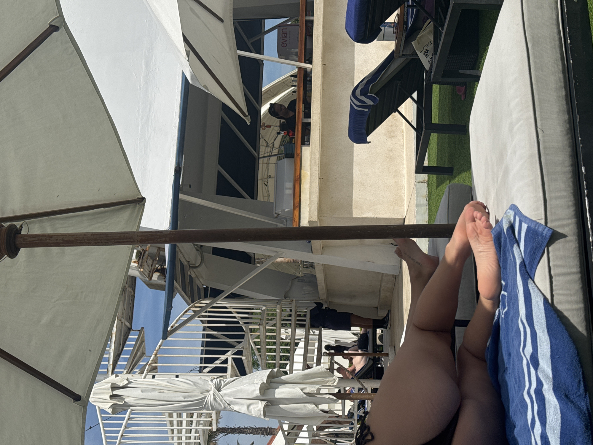 The image, taken horizontally, shows a scene at a beachside location, potentially a resort in North Kuta, Indonesia. A person is lying on a gray-cushioned chaise lounge. Their legs and feet are visible, with a blue and white striped towel partially covering them. To the left, several large white beach umbrellas provide shade. In the background, there is a multi-level building with white railings, indicating a resort or hotel structure. People can be seen on different levels of the building. One person, partially visible, is working behind a bar with an "Evian" sign. The overall environment suggests a sunny day at a resort or beach club in a tropical setting.