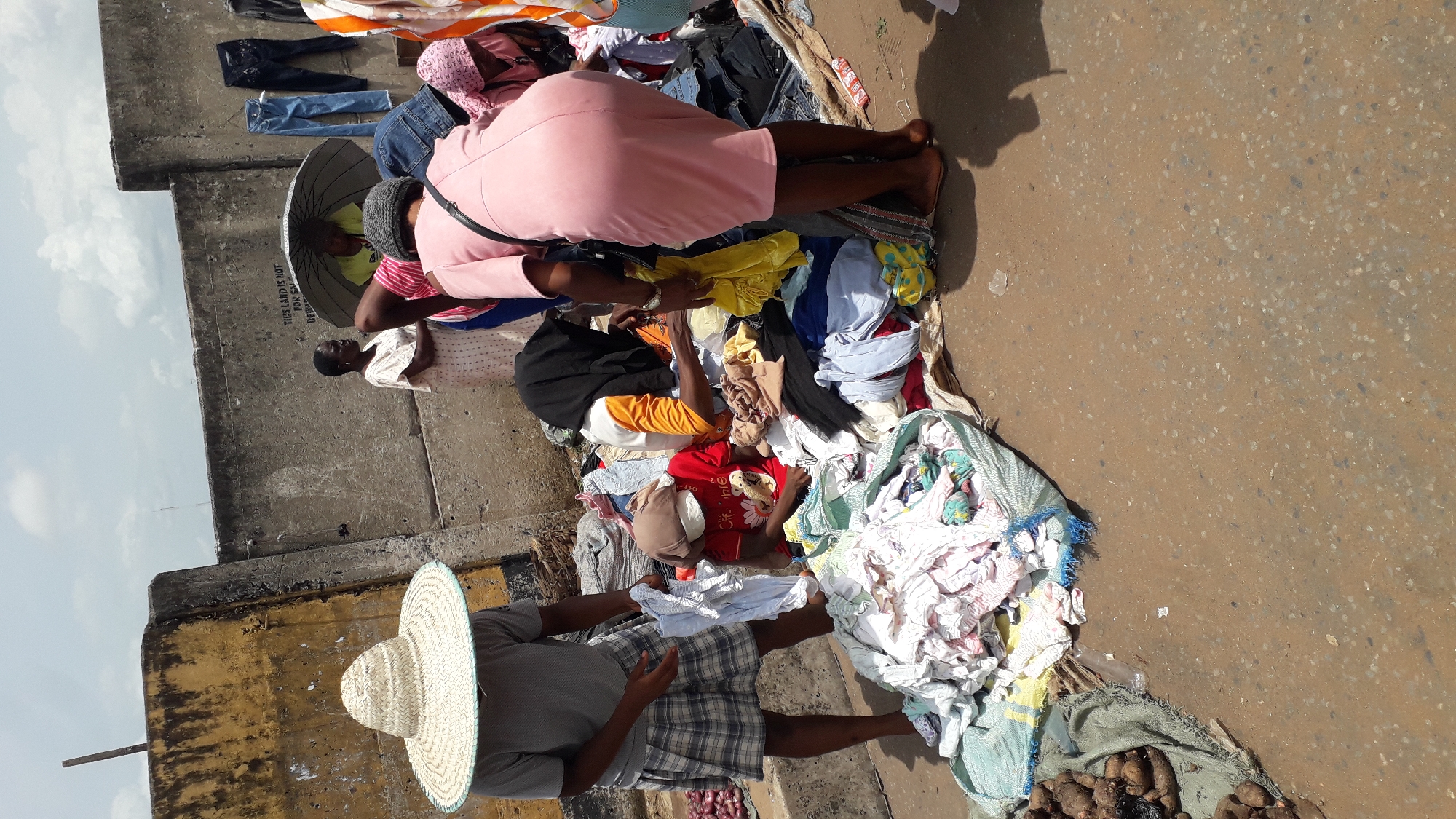 In Onitsha, Nigeria, a street scene depicts several people engaged in commerce. A vendor wearing a large straw hat and plaid shorts stands beside bags of what appear to be sweet potatoes. A pile of colorful clothes is spread out on the ground, with customers inspecting the merchandise. A woman in a pink dress bends over the clothing. Another woman sits under an umbrella. The surrounding area includes a concrete wall with graffiti or markings.