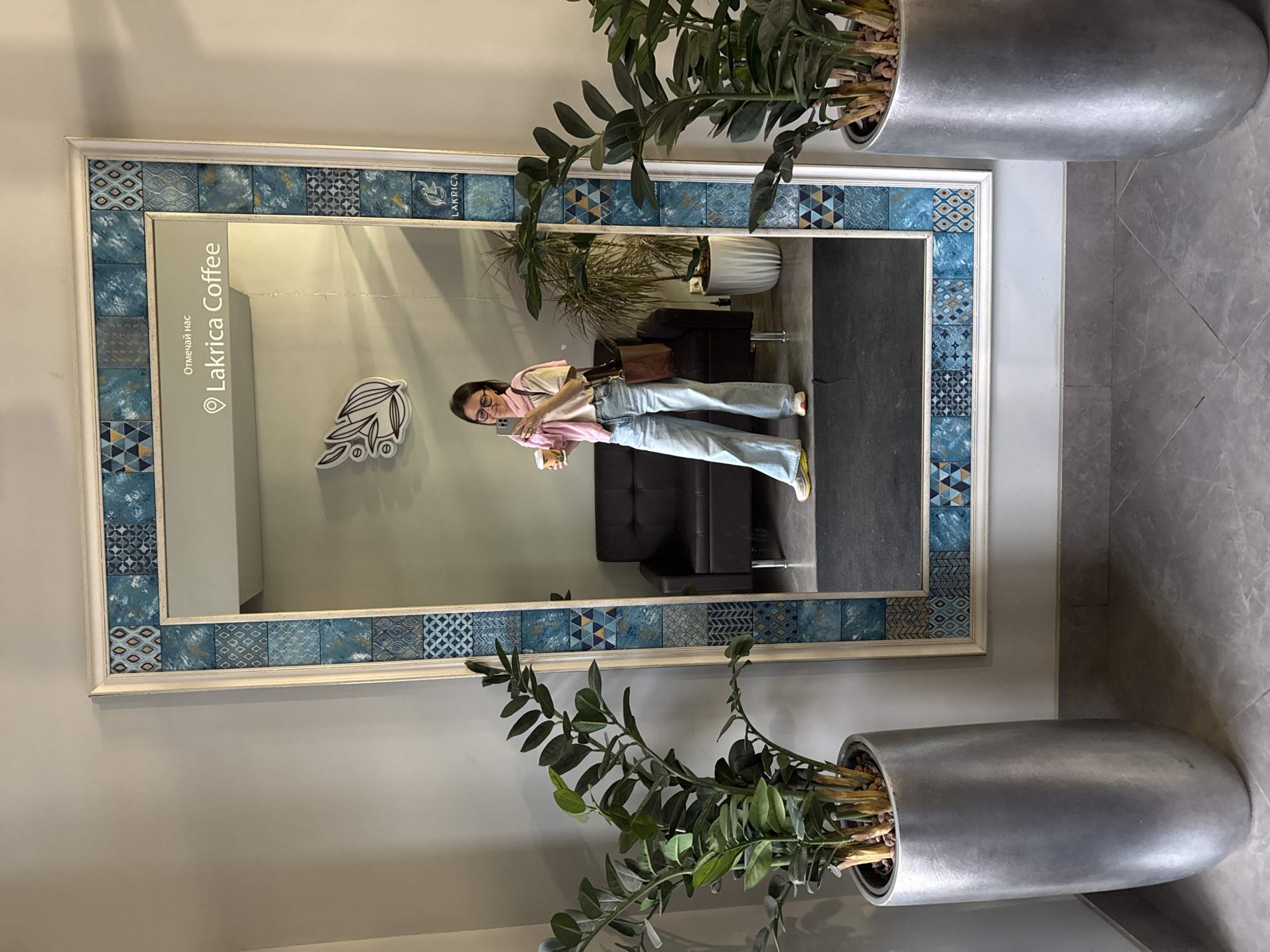 A full-length image shows a woman reflected in a mirror, inside the "Lakrica Coffee" cafe in Izhevsk, Russia. The mirror has a white frame with blue patterned tiles. The woman is standing, holding a phone in her left hand and a cup in her right. She is wearing jeans, a light-colored shirt, and a pink scarf draped over her shoulders. A brown bag hangs off her left shoulder. Behind her, in the mirror's reflection, is a black leather couch. A plant in a white pot is also visible in the background. Two potted plants flank the mirror in the foreground. The floor appears to be grey tile. The wall behind the mirror is a pale grey. "Lakrica Coffee" is written in the reflection on the left side of the mirror.