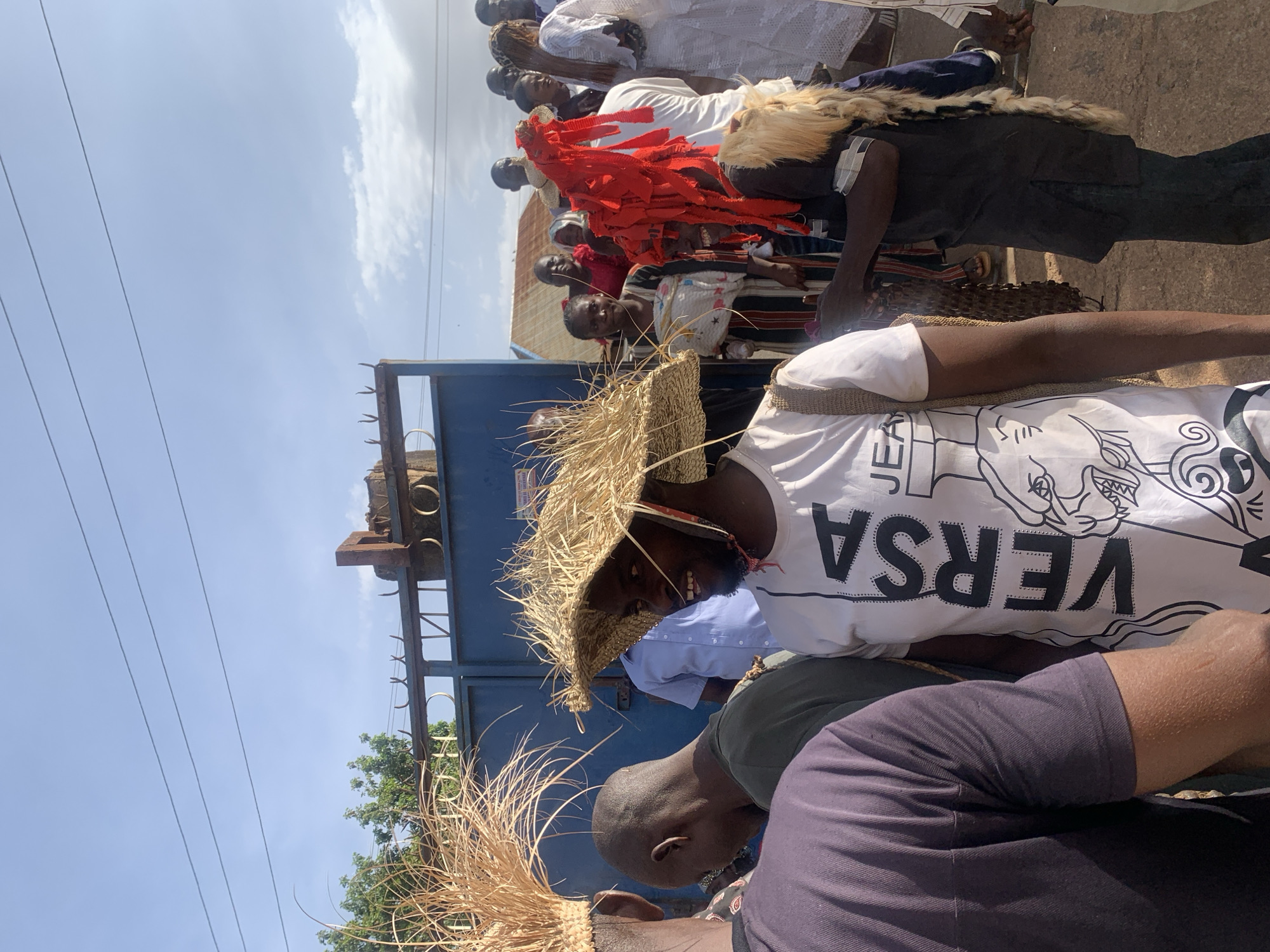 A medium shot shows a group of people lined up on a street in Jos, Nigeria, on a sunny day. The group is diverse in terms of clothing and headwear. Some individuals wear traditional straw hats, while others have on garments with red fabric strips or animal skin details. One person wears a white shirt with the word "VERSA" in bold black letters. Behind them is a dark blue metal gate, and overhead are electrical wires against a clear sky.