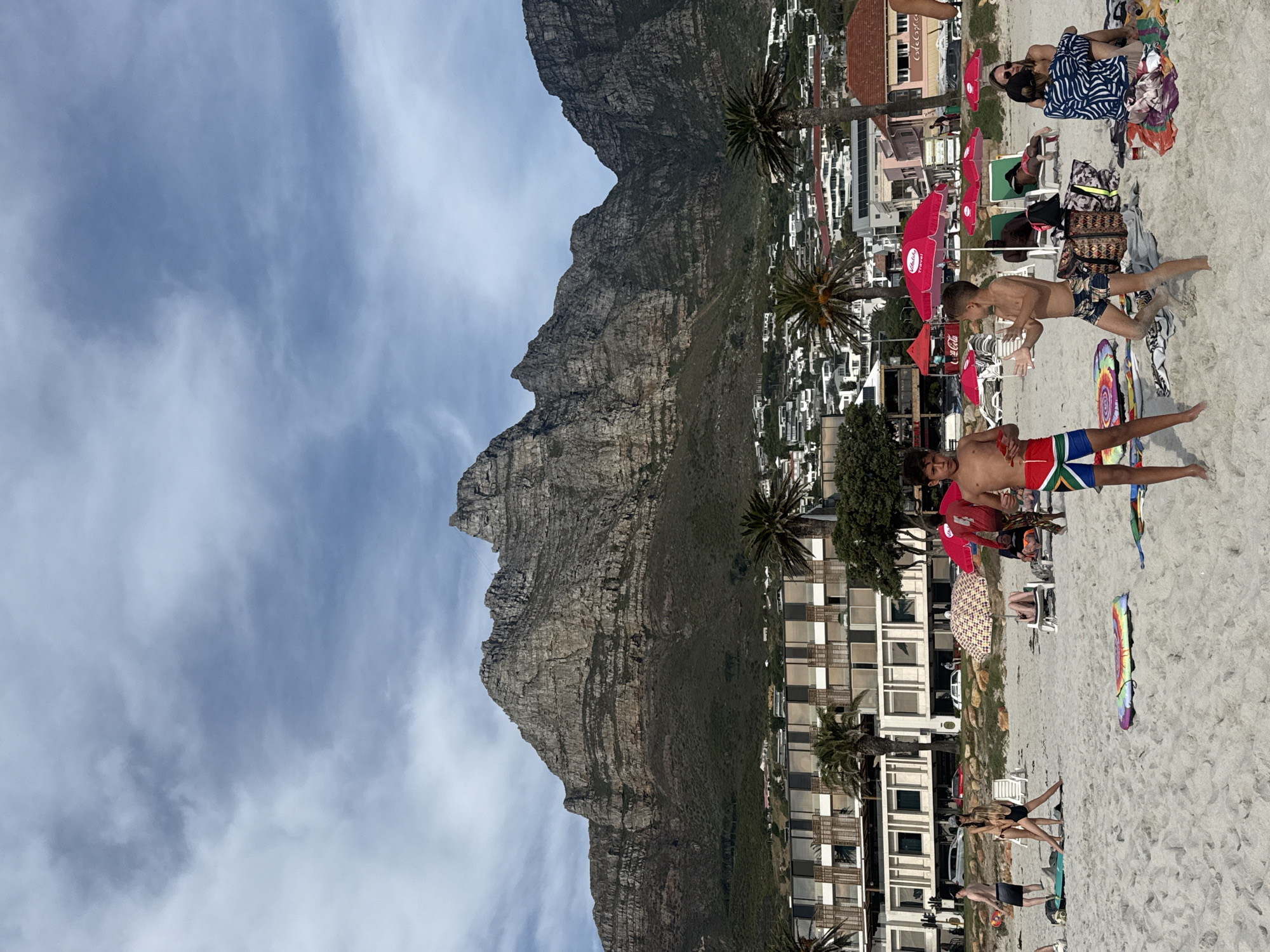 The image captures a scene at Camps Bay beach in Cape Town, South Africa, with the distinctive silhouette of Table Mountain in the background.
In the foreground is the sandy beach, where various individuals are engaged in leisure activities. Several people are sunbathing on towels, some under red beach umbrellas bearing the Coca-Cola logo. Two boys are standing near the water's edge; one is wearing swim trunks bearing the South African flag. A few buildings and palm trees are adjacent to the beach. The sky above the mountain is partly cloudy.