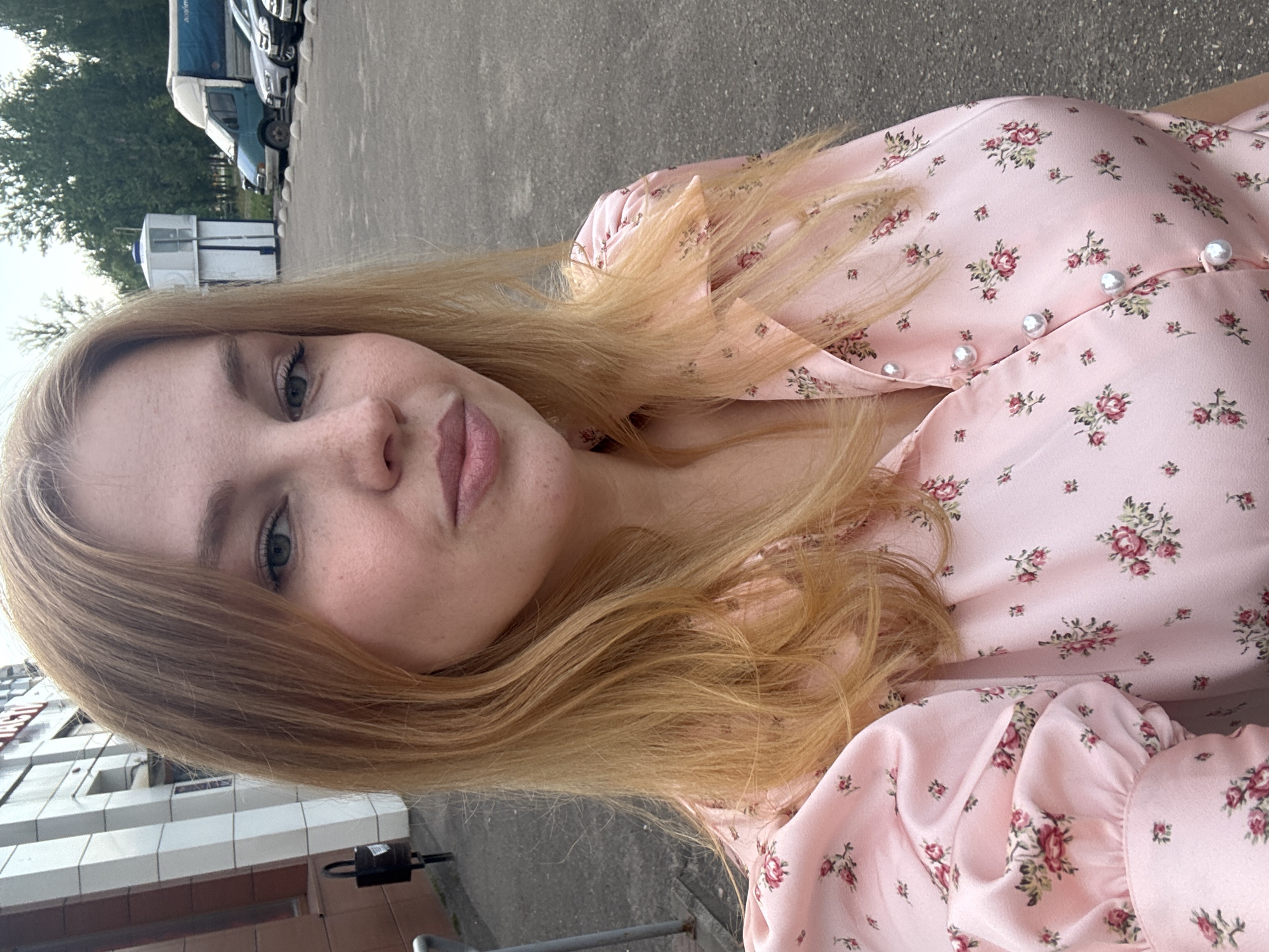 In Yaroslavl, Russia, a blonde woman with fair skin, freckles, and light green eyes is pictured outdoors. She is wearing a pink satin blouse with a floral pattern and pearl-like buttons. The background shows an asphalt parking lot with several cars parked, including a blue and white truck, and a building with a beige and white facade. Trees are visible in the distance. The lighting suggests it is daytime.