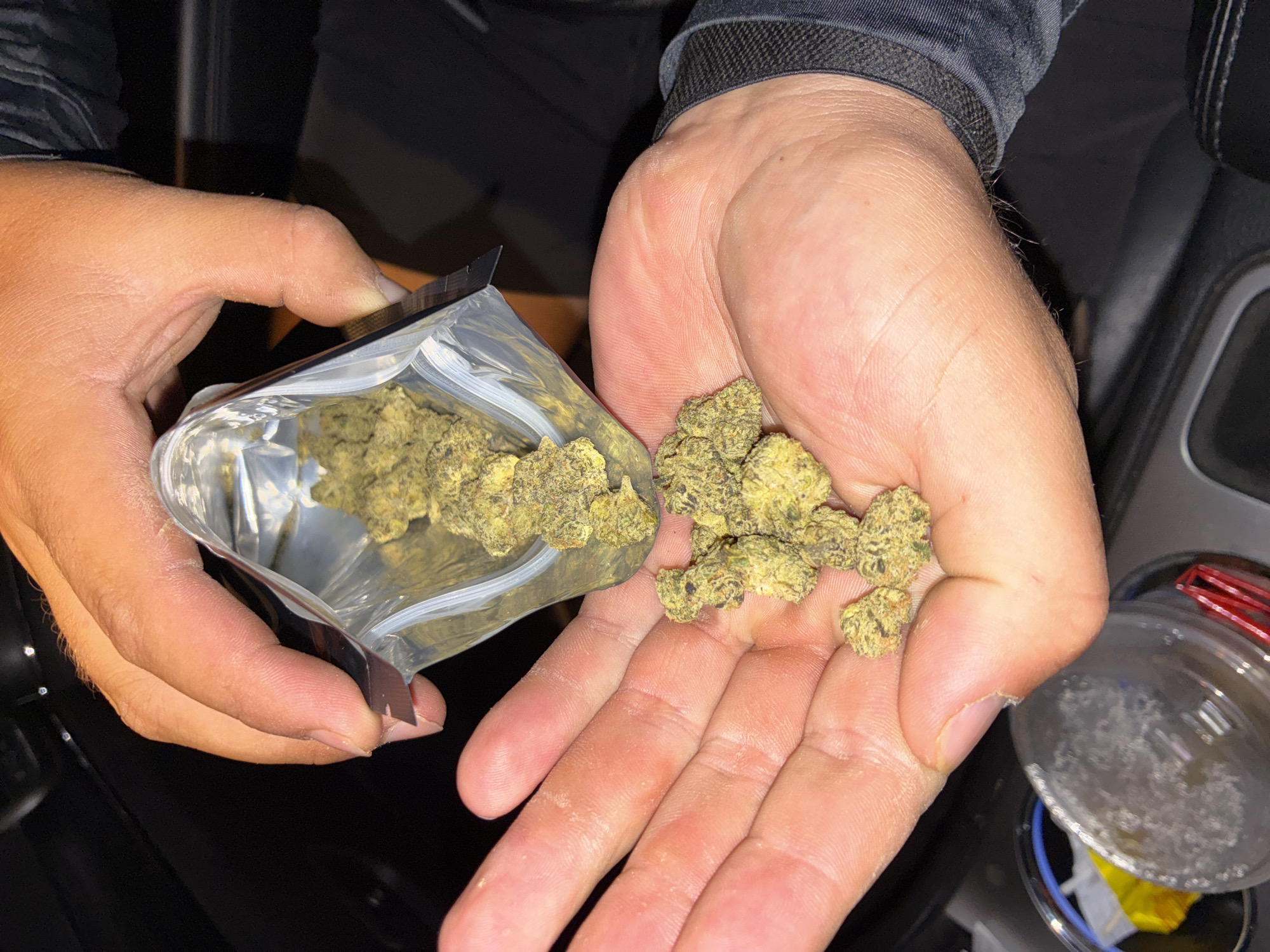 The image presents a close-up view of two hands handling green, leafy material, strongly resembling cannabis buds. The left hand holds a clear, open, resealable plastic baggie, from which more of the same material is visible. The right hand cups a small pile of these green buds, suggesting a transfer or measurement is taking place.
The setting appears to be inside a vehicle, with parts of a car's dark interior, possibly a center console, visible in the background. In the lower right corner, a clear plastic cup with some white residue and a red clip on its rim sits within a cupholder, reinforcing the car interior location. The lighting is dim, indicating it might be nighttime or an indoor, poorly lit environment.
The hands are adult, showing visible skin texture and creases. No faces are visible, so no emotions can be determined. There is no visible text on the baggie, hands, or other objects in the frame. The overall activity depicted is the handling and apparent transfer of what is likely marijuana from its packaging into a person's palm. The provided location context is Huixquilucan de Degollado, Mexico.