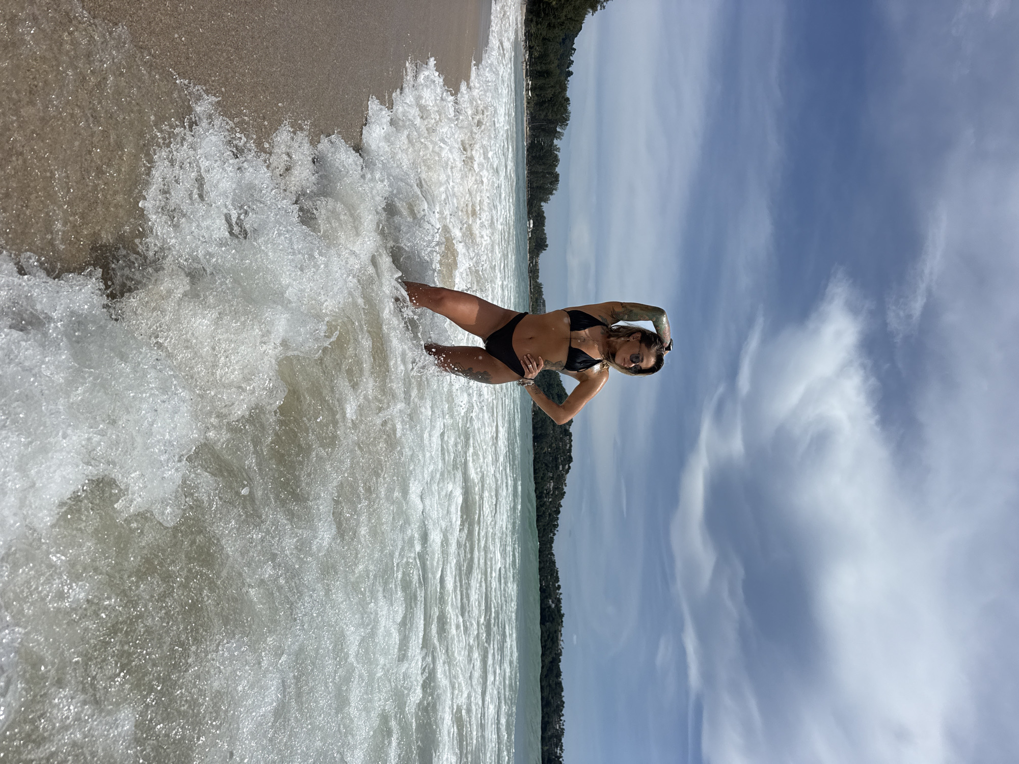 A woman stands in the shallow ocean water on a beach, wearing a black bikini. She has tattoos on her arms and is looking to her right with one hand on her hip and the other raised behind her head. The ocean waves are breaking around her, creating a foamy surf. The sand is visible to her left, and the sea stretches out before her, meeting a distant, tree-covered shoreline under a partly cloudy sky. The location is specified as Choeng Thale, Thailand.