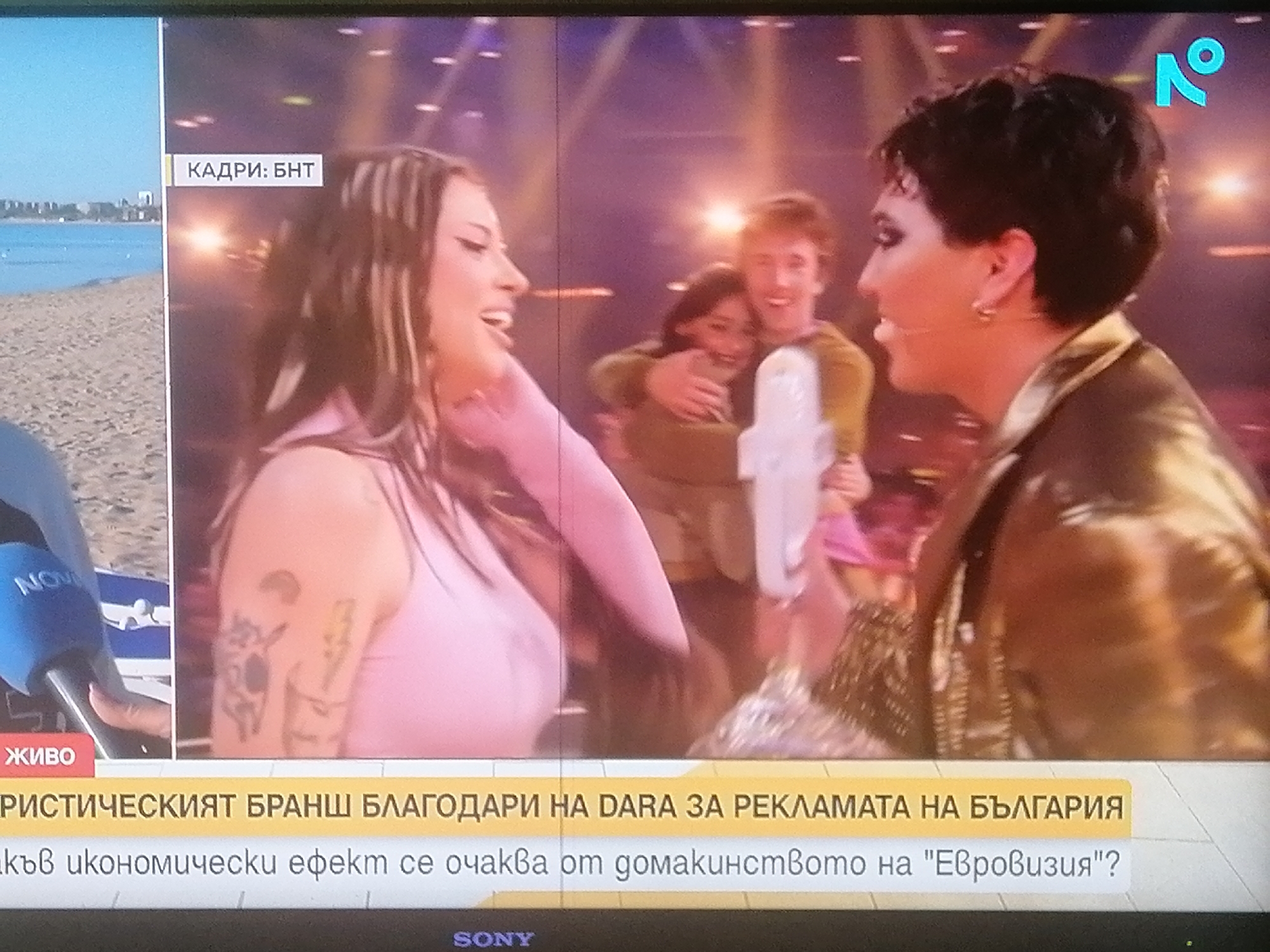 The image appears to be a screen capture of a TV broadcast, likely news, showing two separate images: One image is a beach scene with buildings in the background, likely in Sofia, Bulgaria. The second image is an indoor scene with several people. On the left side, a woman with long hair, tattoos on her arm, and wearing a pink top is shown in profile, smiling and seemingly talking to someone off-screen. On the right, a person with short, dark hair and wearing a shiny jacket is also in profile, likely engaged in the same conversation. Behind them, another person is hugging a young girl. There is text at the bottom of the screen in Bulgarian, mentioning "Dara" and "Bulgaria," suggesting the topic may be about her advertising for Bulgaria. A microphone with a blue logo is visible on the left, near the beach scene. A logo is visible in the upper right corner. The TV screen also displays "КАДРИ: БНТ" (Frames: BNT), likely a Bulgarian TV station. The bottom of the screen features the Sony logo.