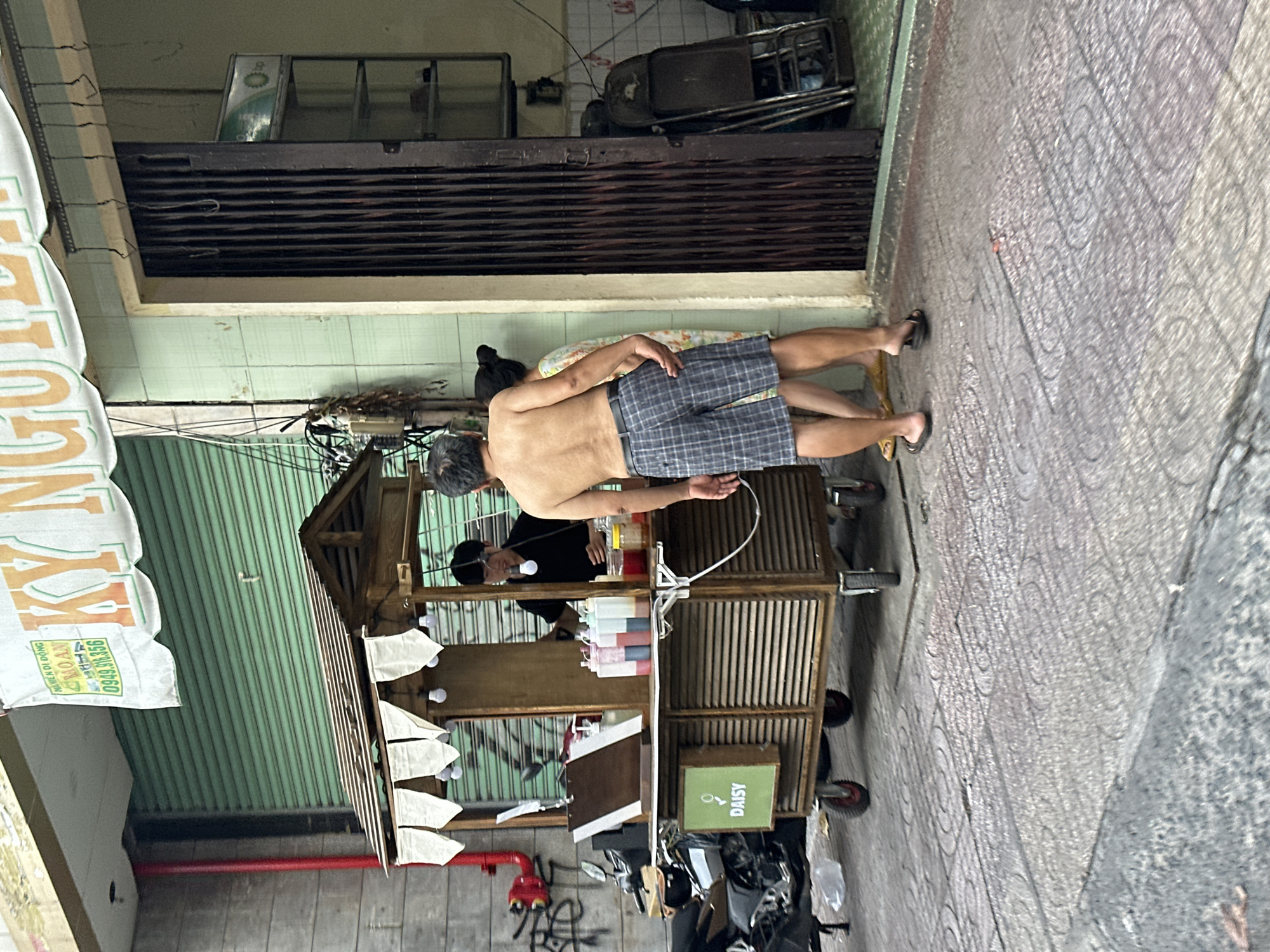 Thủ Đức, Vietnam: A man with grey hair and a bare torso wearing plaid shorts stands at a wooden street cart vending beverages. A person in a black shirt stands behind the cart. A woman lies on a patterned cloth nearby. A closed, dark metal gate is above the scene. A motorcycle is parked near the cart. The street is paved.