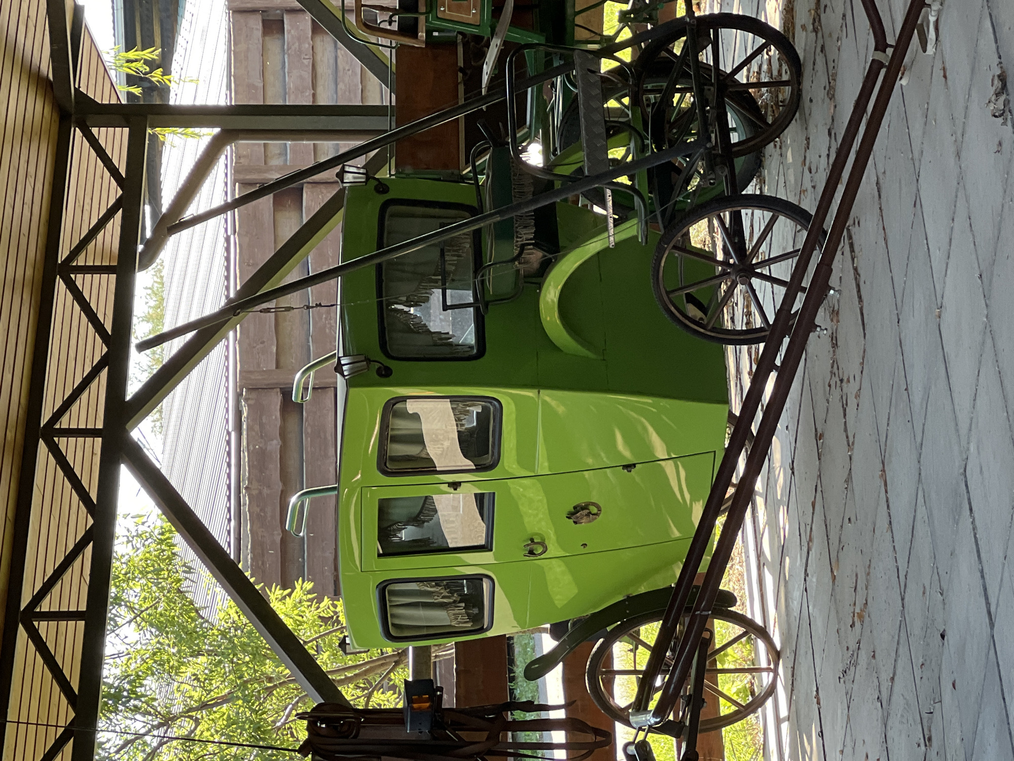 A bright green horse-drawn carriage is parked under a wooden and metal roof structure. The carriage has four windows with draped curtains and two lanterns attached to the exterior. It is supported by two large spoked wheels at the rear and two smaller spoked wheels at the front. In front of the carriage, a metal frame with a seat and more spoked wheels suggests another, smaller carriage or vehicle. The ground is paved with square tiles. In the background, a wooden fence and a glimpse of trees and a building can be seen. The scene appears to be outdoors, possibly in a rural or historical setting. There are no people present in the image.