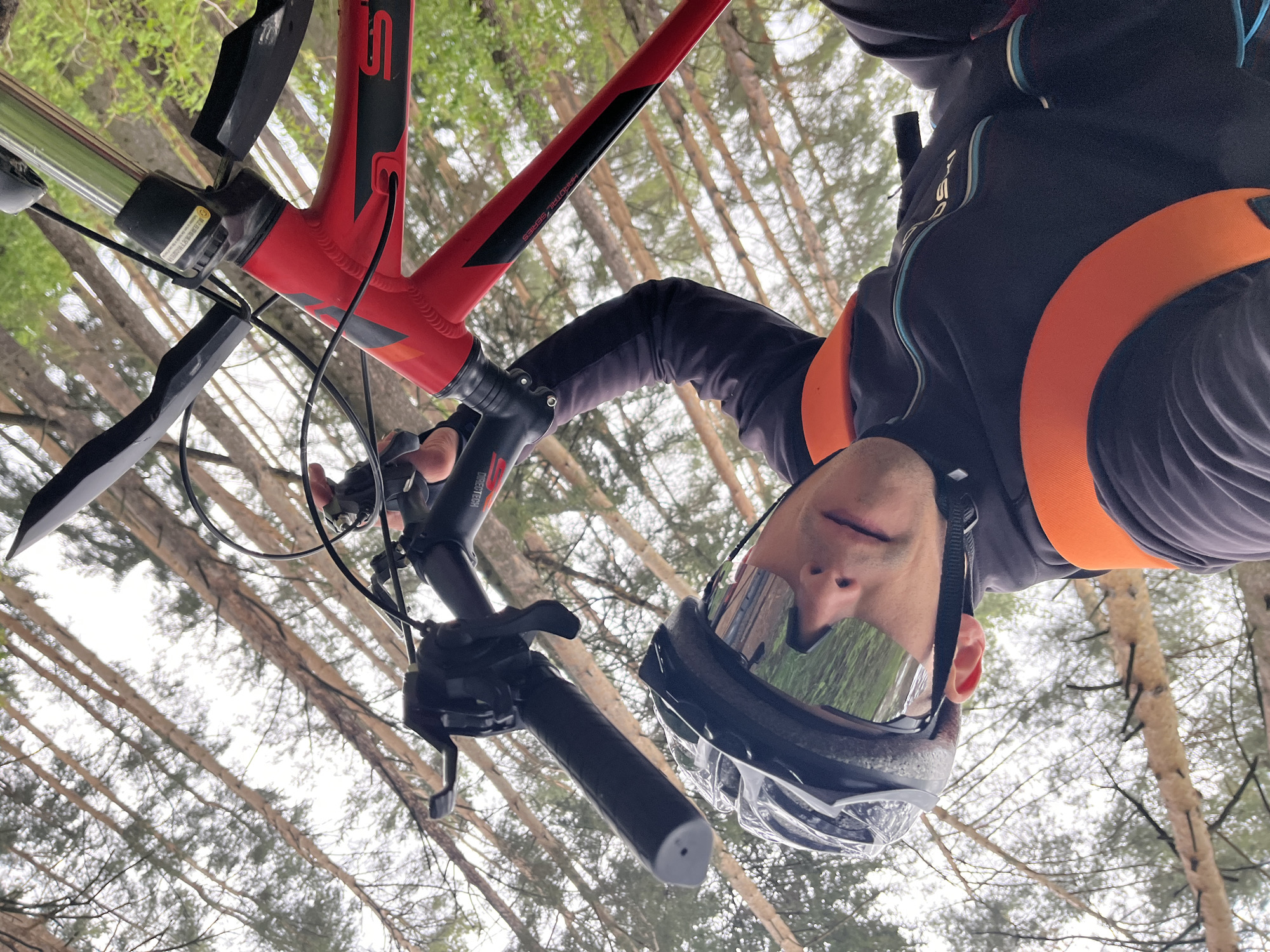 The image shows a person on a red mountain bike, possibly in a wooded area near Chaikovsky, Russia. The person is wearing a black helmet with mirrored goggles and a dark jacket with orange trim. The camera is positioned looking upwards at the cyclist, capturing the bicycle's handlebars and frame against a backdrop of tall, thin trees. The scene appears to be outdoors, likely in a forest or park setting. The specific location in Chaikovsky cannot be determined from the visual information provided.