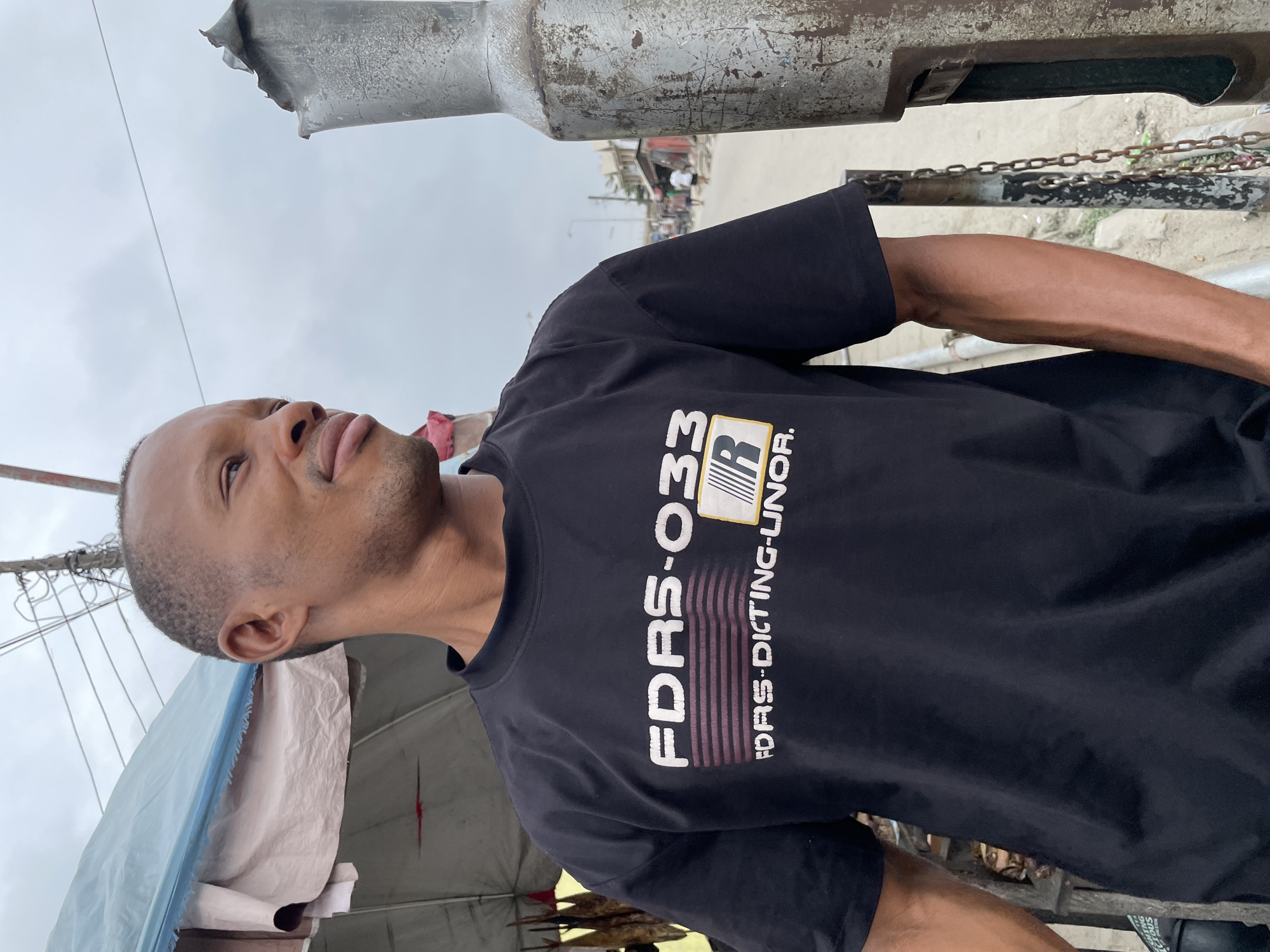 A man in a black t-shirt with white text that reads "FDRS-033" and "FDRS-DICTING-UNOR." stands outdoors in a slightly overcast environment, possibly in Ado, Nigeria. He is looking to his right with a slight smile. Behind him, a blue and tan tarp covers what appears to be a market stall or shelter. The man's left arm is visible, and to his right, a rusty metal pole with a chain wrapped around it is prominent. In the background, a dusty street with buildings and a few people walking can be seen, suggesting an urban or semi-urban setting.