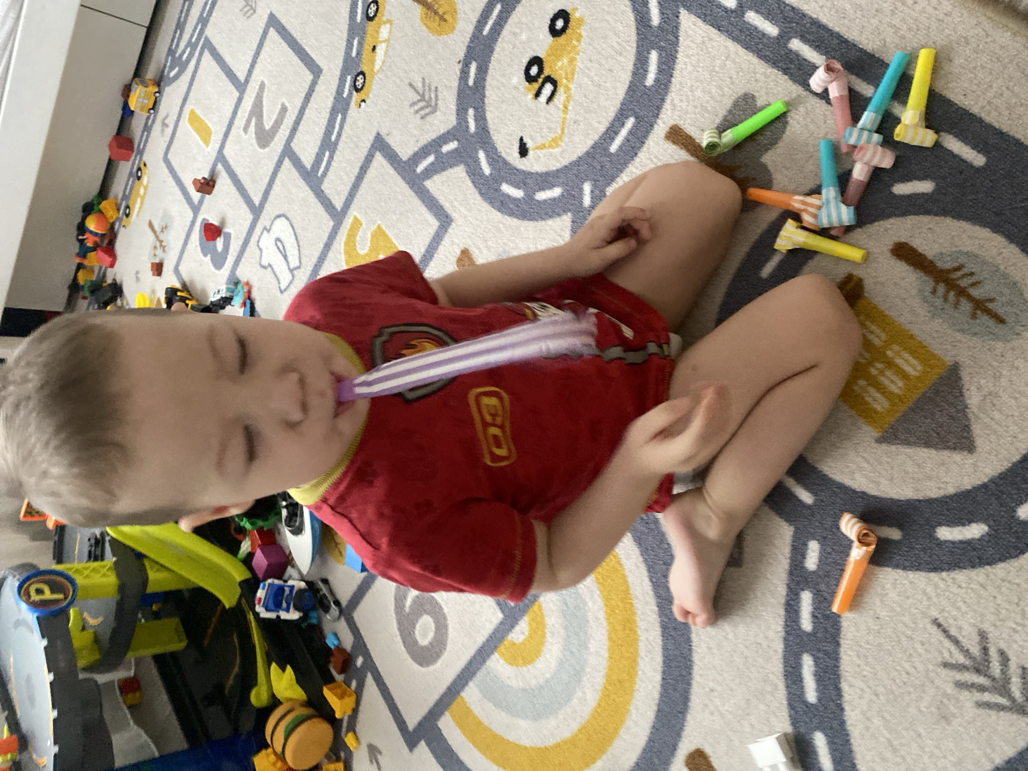 This image captures a young child sitting on a themed rug in what is identified as Sochi, Russia. The child, with light hair, is wearing a red short-sleeved shirt with a yellow emblem on the front and green trim, along with a light-colored undergarment, and is barefoot. The child is actively blowing into a purple and white striped party blower, causing it to extend. The child's eyes are partially closed.
The setting is an indoor space, likely a child's play area, characterized by a light-colored rug featuring a road and car pattern with various shapes and numbers like "2" and "3". Numerous toys are scattered across the floor in the background and foreground. Visible toys include a multi-level yellow and grey toy parking garage with a "P" sign and a green ramp, various toy vehicles such as a blue and white police car, a grey car, and a yellow bus. Other toys consist of colorful building blocks (red, yellow, orange, blue, purple), a large round yellow toy resembling a hamburger bun, and a white toy boat. To the child's right, a collection of unblown party blowers in various colors including orange, yellow, light blue, pink, green, and light brown are dispersed on the rug. A white piece of furniture is partially visible along the right edge of the background.