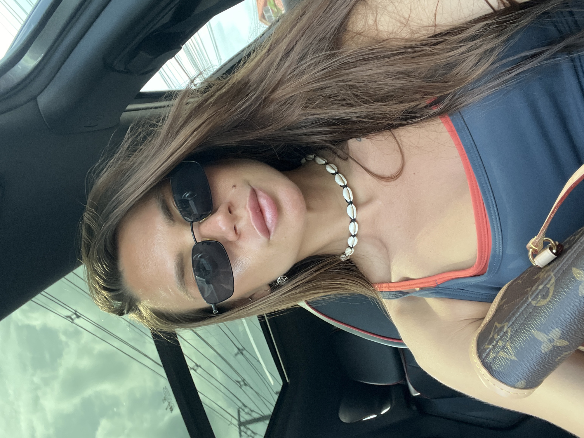 A woman with long, brown hair, wearing sunglasses and a shell necklace, sits in a car. She is holding a brown designer wallet. The car has a sunroof, through which the sky and power lines are visible. The scene appears to be in a city, possibly Baan Bo Phut, Thailand, given the greenery and architecture visible outside the car.