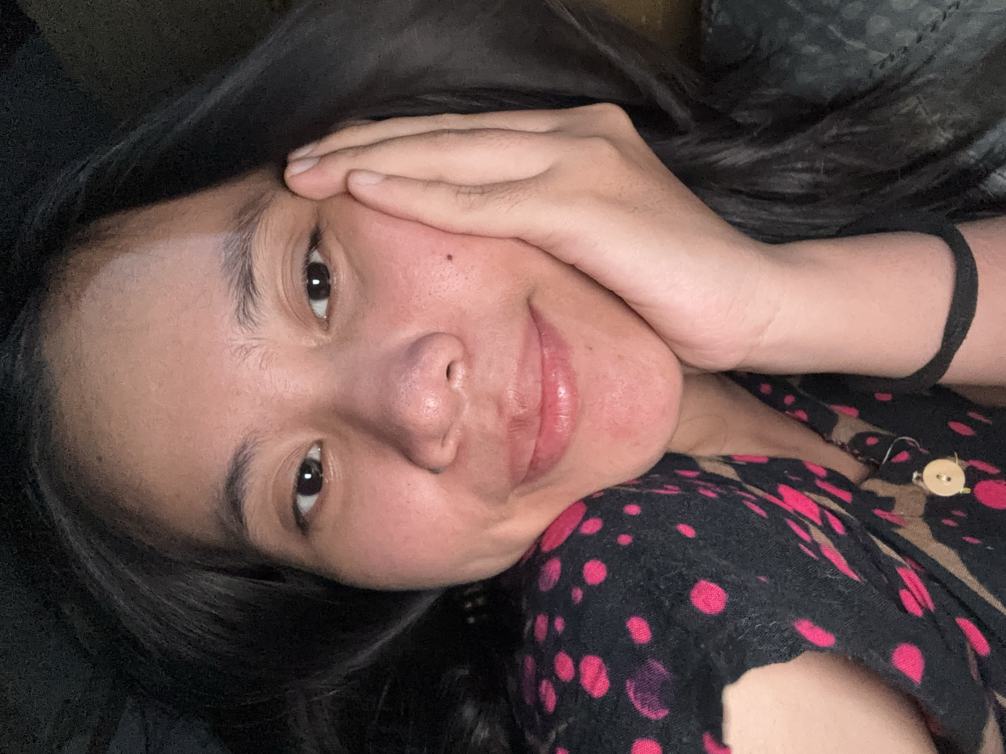 A young woman with dark hair is looking at the camera, resting her right cheek on her hand. She is wearing a black shirt with pink polka dots and a brown and tan pattern. The background is dark, suggesting the photo was taken indoors at night. The image does not contain any information to determine the location in the city of East Jakarta, Indonesia.