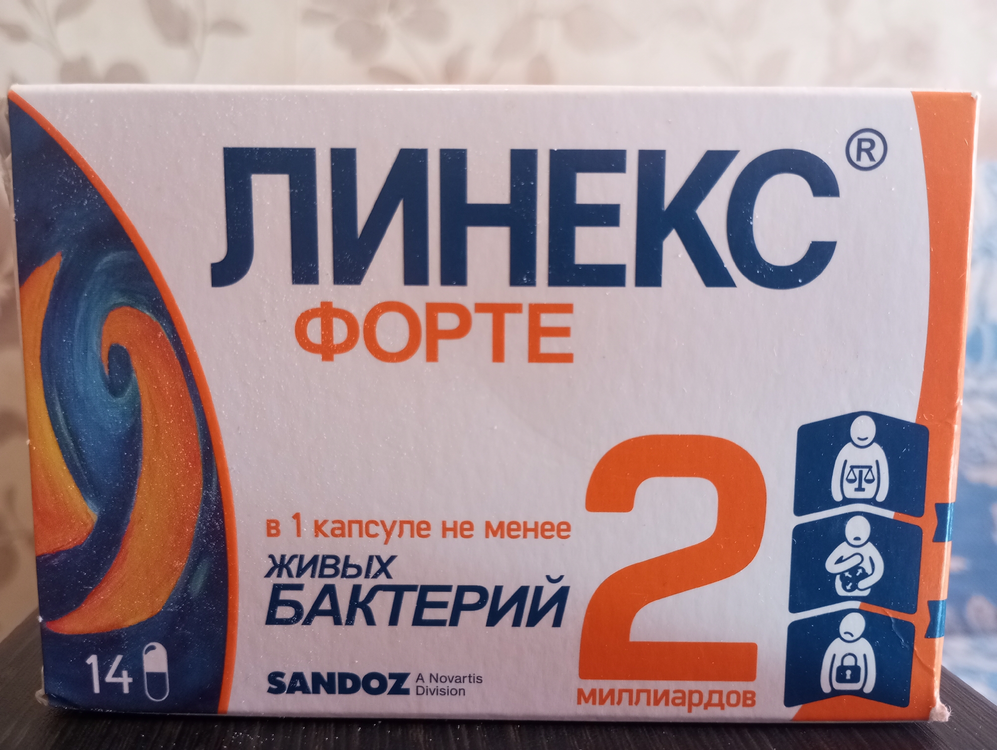 The image shows a close-up of a box of "Linex Forte" medication. The box is primarily white with dark blue and orange lettering. The brand name "ЛИНЕКС" is prominently displayed in large dark blue capital letters, with "ФОРТЕ" in smaller orange capital letters below it. To the left, there is a stylized orange and blue swirl design. The bottom left corner of the box shows the number "14" followed by an icon of a capsule. Text in Russian on the box reads "в 1 капсуле не менее ЖИВЫХ БАКТЕРИЙ" which translates to "in 1 capsule not less than LIVE BACTERIA." Further to the right, a large orange number "2" is visible, with the word "МИЛЛИАРДОВ" (billions) below it. On the right side of the box, there are three blue rectangular icons, each with a white silhouette of a person and a symbol. The top icon shows a person with scales, the middle shows a person holding a stomach, and the bottom shows a person with a lock. The brand "SANDOZ" is visible in the lower left quadrant, with "A Novartis Division" below it. The image appears to be taken indoors, with a textured, light-colored background visible behind the box. The box is placed on a dark, possibly wooden surface. There are no people visible in the image. The location in the city of Kirzhach, Russia, is not determinable from the provided image.