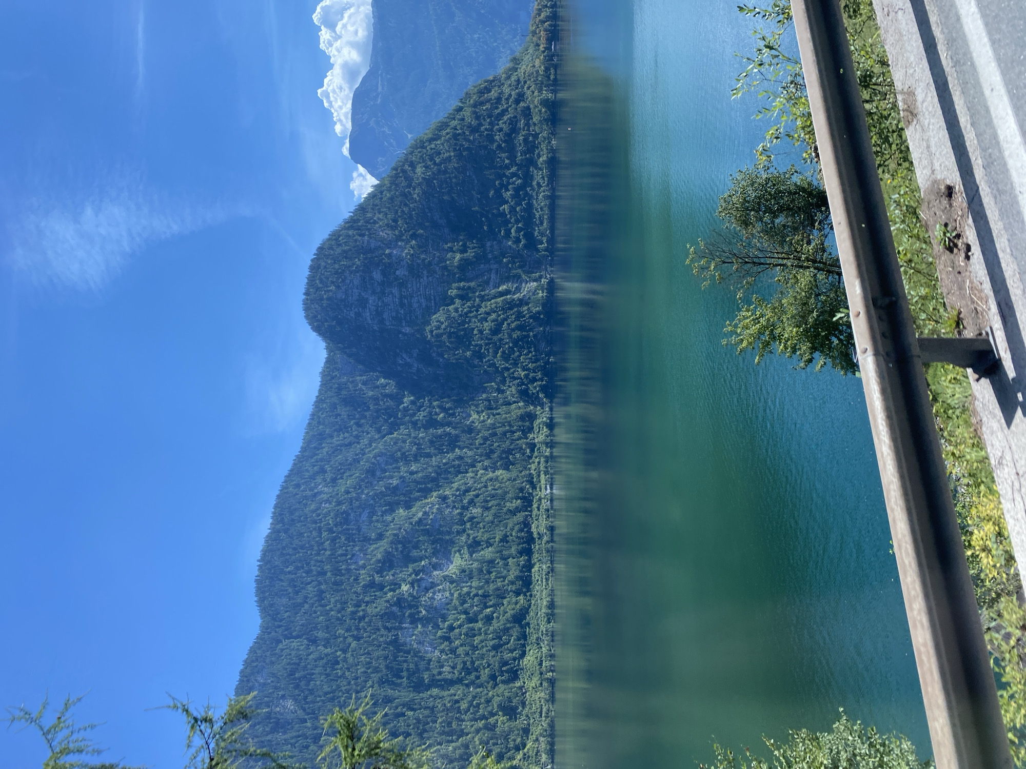 A wide-angle, eye-level shot captures a serene lakeside scene in Hallstatt, Austria, on a clear, sunny day. The foreground features a guardrail along a paved road, with sparse green vegetation growing alongside it. Beyond the road, a vast expanse of calm, deep teal water dominates the lower half of the image. The water's surface reflects the surrounding landscape, creating a mirror-like effect.
In the mid-ground, a dense, dark green forest blankets the steep slopes of a massive, rocky mountain that rises dramatically from the water's edge. The mountain's surface is textured with visible rock formations and is densely packed with trees, giving it a rugged, imposing appearance. A thin, lighter green band of vegetation runs along the base of the mountain where it meets the lake.
The background is characterized by a bright, clear blue sky with a few wispy white clouds. To the right, distant, hazy blue mountains with white, puffy clouds are visible. A single, small white object, possibly a boat, is discernible on the water in the mid-ground, near the treeline of the mountain. There are no people or animals visible in the scene. The overall impression is one of natural beauty and tranquility.