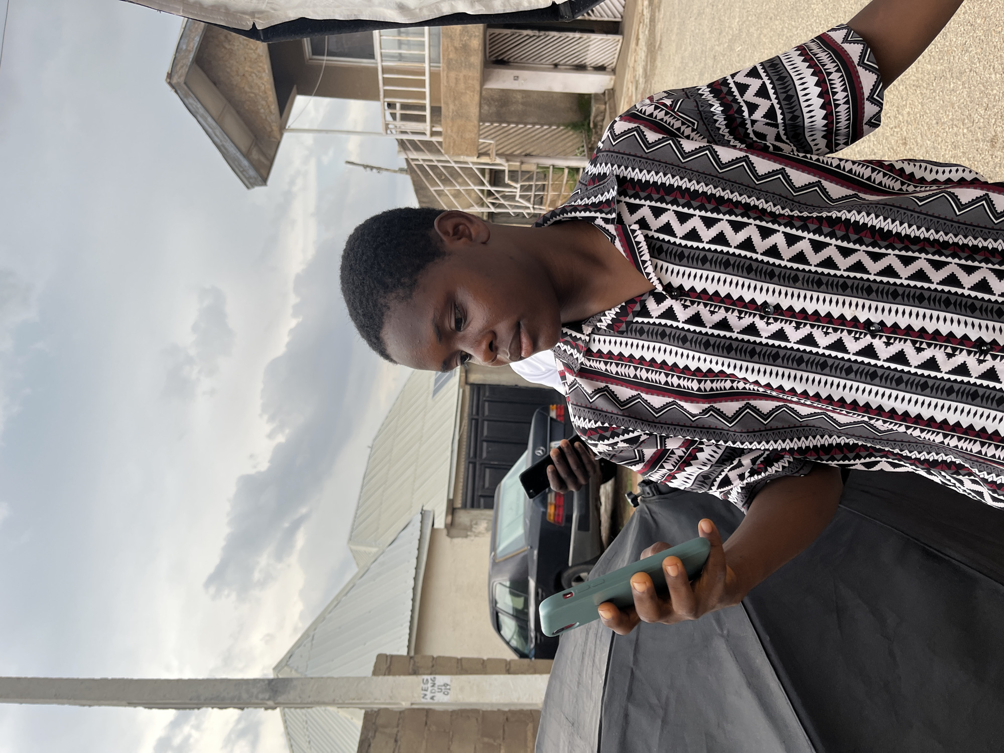 The image features a person in the foreground holding a mobile phone. They are wearing a patterned shirt with geometric designs in black, white, and red. The background shows buildings, one with multiple floors and balconies, and a black car parked in front of a garage door. The sky is overcast with clouds, suggesting a cloudy day. The scene appears to be an outdoor setting, possibly a residential or commercial area. There is no visible text in the image.