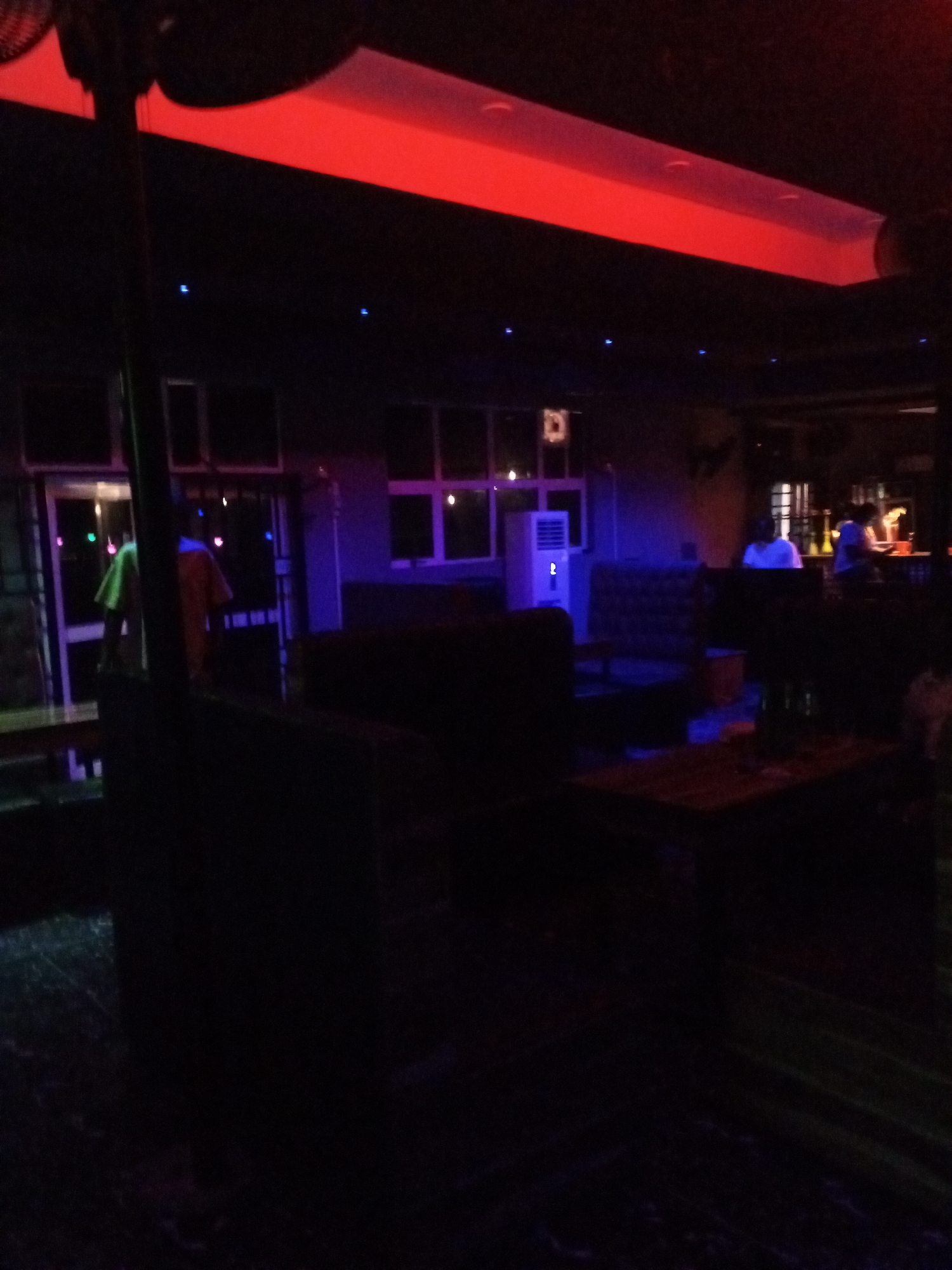 The image shows the interior of a dimly lit establishment, possibly a bar or lounge, with red and blue lighting. In the background, there are tables and seating areas. Several people are visible, some standing and some seated, though their features are not clearly discernible due to the low light. One person in the foreground appears to be wearing a yellow garment. Another individual is seen in an orange shirt. In the far background, some bottles are visible on a counter. The overall ambiance is that of a nighttime venue.