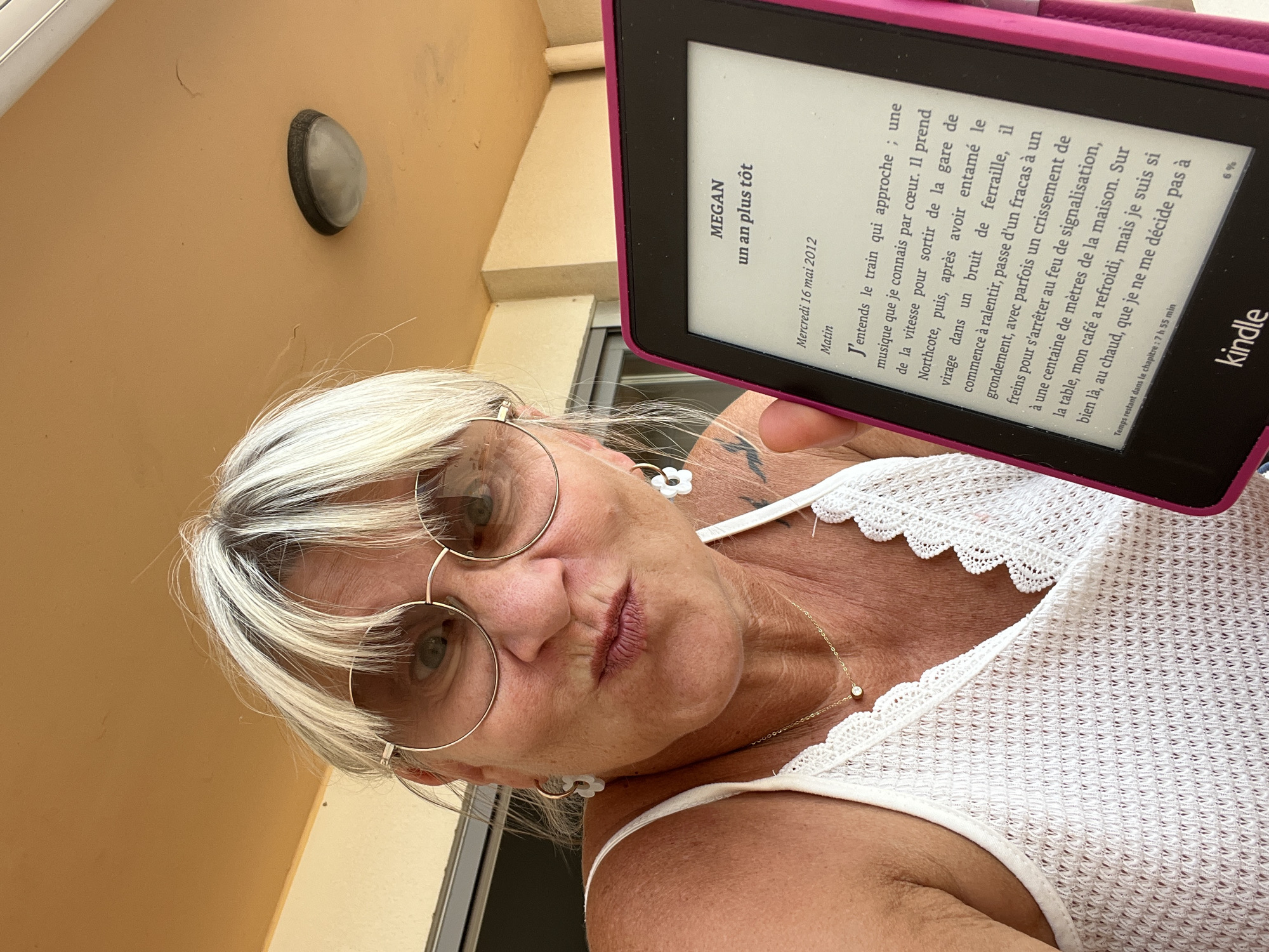 A woman with blonde hair and bangs, wearing round glasses and a white lace tank top, holds up a pink Kindle e-reader. She is making a kissing face towards the camera. The Kindle displays text in French, with the title "MEGAN un an plus tôt" visible. The background shows a light-colored wall and ceiling with an outdoor light fixture, suggesting an outdoor covered area, potentially a balcony or terrace. The scene is likely captured in Palavas-les-Flots, France, given the context of the surrounding area.