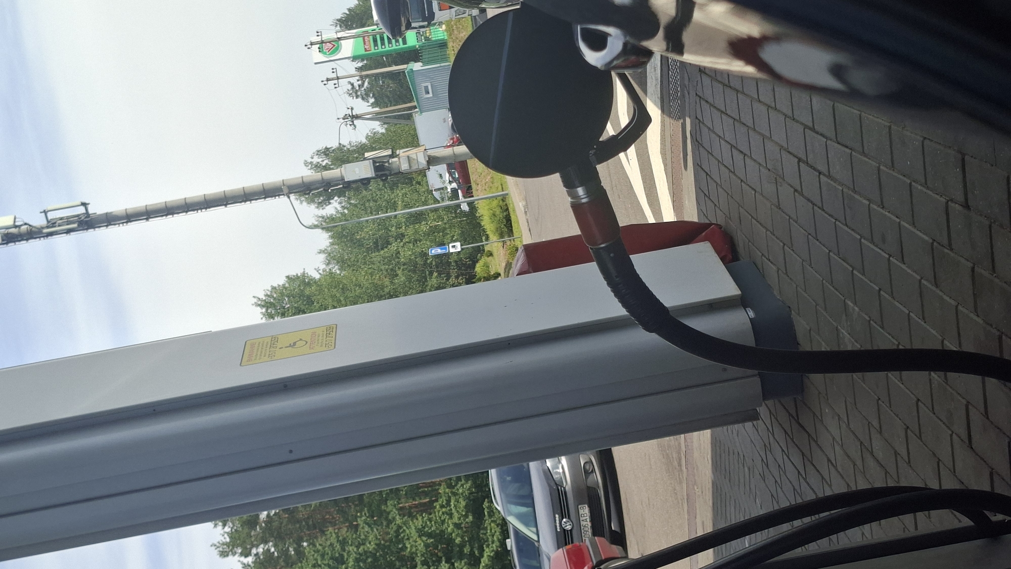 An outdoor shot captures a gas pump nozzle connected to a car's fuel filler on a sunny day. The scene is set at a gas station, possibly in the Драчкаўскі сельскі Савет, Belarus, as indicated by the Cyrillic text on a yellow sticker on the gas pump which reads "Внимание" (Attention). The sticker also contains a phone number: "+375 17 2093539" and a wheelchair symbol, suggesting accessibility information.
In the background, trees and a cell tower are visible. A dark-colored Volkswagen station wagon is parked nearby, displaying a license plate "0906 AB-8". The ground is paved with interlocking gray bricks. The composition is framed by the gas pump column on the left and the car's side mirror and fuel cap area on the right. The overall lighting suggests daytime with clear skies.