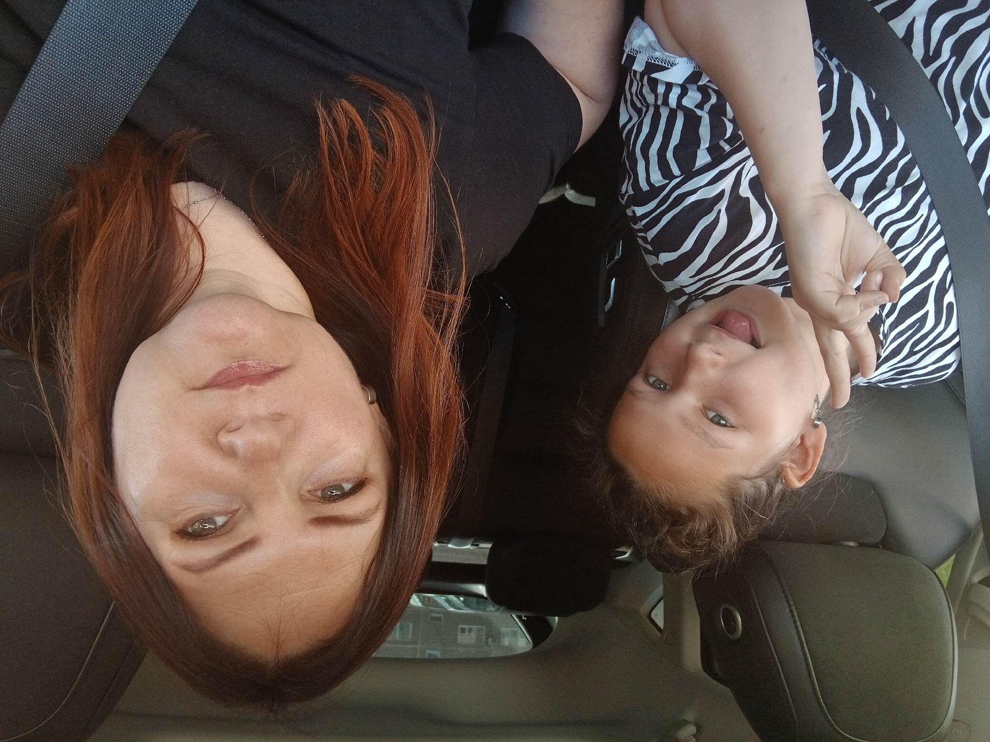 Two females, an adult and a child, are seated in a car. The adult, with reddish-brown hair, is in the foreground on the right. The child, with light brown hair pulled up in a bun, is on the left, wearing a black and white striped shirt. The child is sticking out their tongue and making a peace sign with their fingers. Both are wearing seatbelts. The car's interior is visible, with dark gray seats and headrests. Outside the car window, buildings and parked cars can be seen, suggesting an urban environment, possibly in Chaikovsky, Russia. The image appears to be a selfie taken from inside the car.