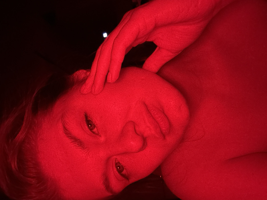 A person's face and hand are bathed in intense red light. The face is tilted, with eyes open and mouth slightly parted. A hand rests on the forehead, fingers extended. The red light obscures details and creates a stark, almost monochromatic image, highlighting the texture of the skin. A dark background adds contrast.