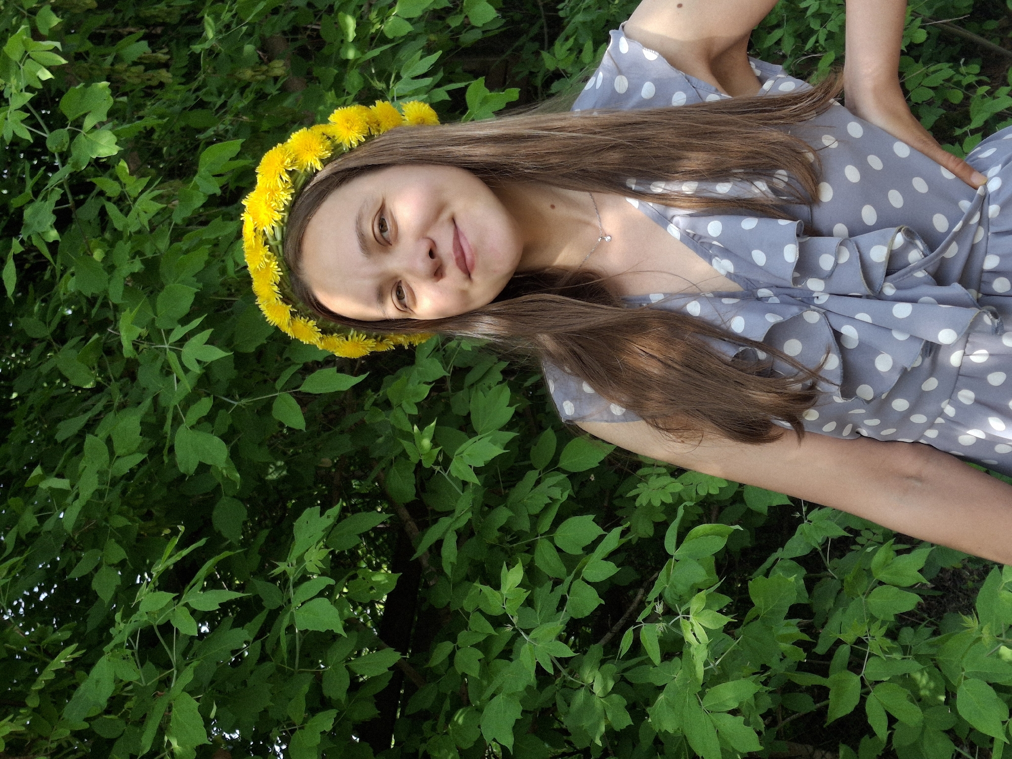 The image shows a young woman wearing a dandelion crown and a grey polka-dot dress, set against a backdrop of lush green foliage. She is smiling at the camera. A silver necklace is visible around her neck. The background suggests a natural setting, possibly a park or wooded area in Chaikovsky, Russia. The location can only be surmised as the request for it to be in the city of Chaikovsky, Russia, but there is no evidence it is necessarily from there.