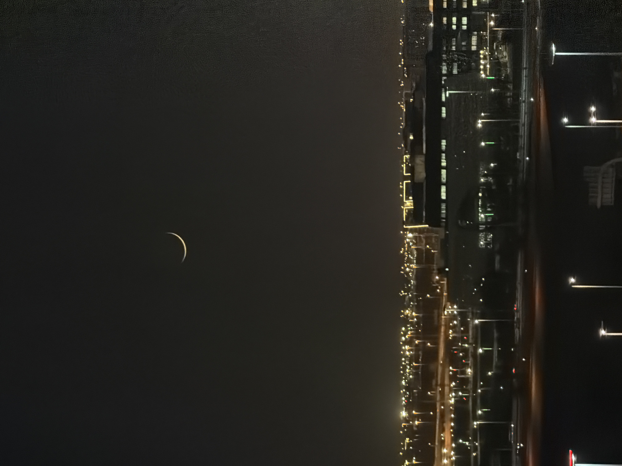 A crescent moon is visible in the dark night sky over an urban landscape. The moon is a bright sliver against the blackness. Below the moon, the city lights are spread out, forming a luminous horizon. Buildings with illuminated windows are visible, along with streetlights that punctuate the darkness. The overall impression is of a city at night, with the celestial body offering a gentle light above.