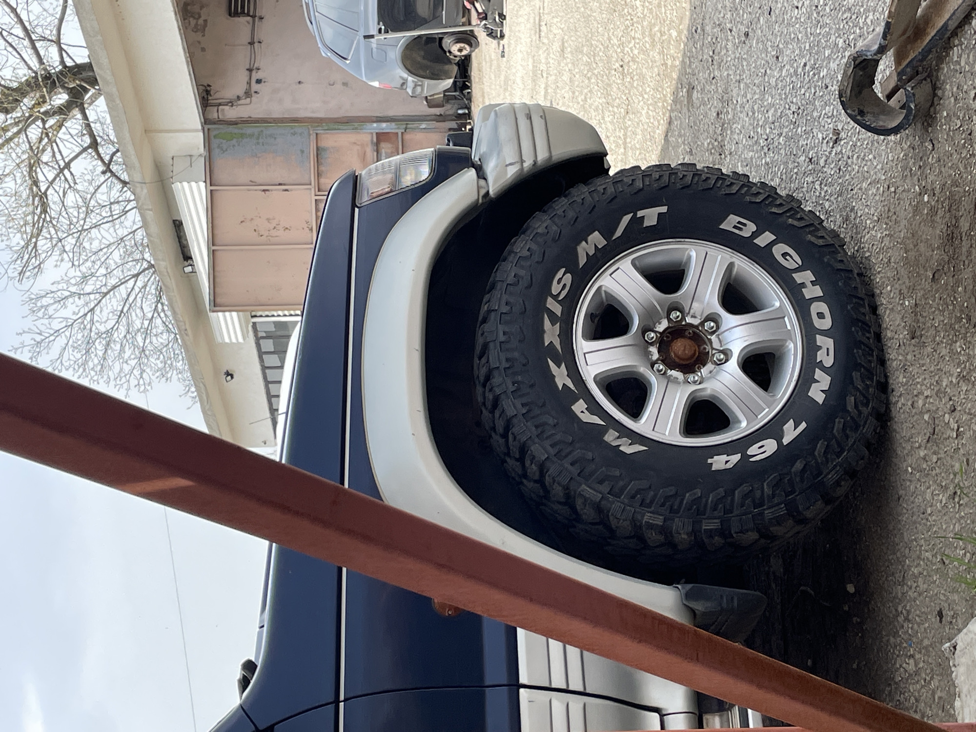 The image shows the front driver's side of a dark blue and white vehicle, likely a sport utility vehicle, parked in what appears to be an outdoor workspace or garage area. The most prominent feature is the large tire with the brand name "BIG HORN" visible on the sidewall. The vehicle is parked near a building with faded pink walls and a white awning. Other vehicles are partially visible in the background. The presence of repair equipment suggests that this location is an automobile shop or repair facility. The image does not provide enough information to confirm a specific location in Vabel, Bulgaria.