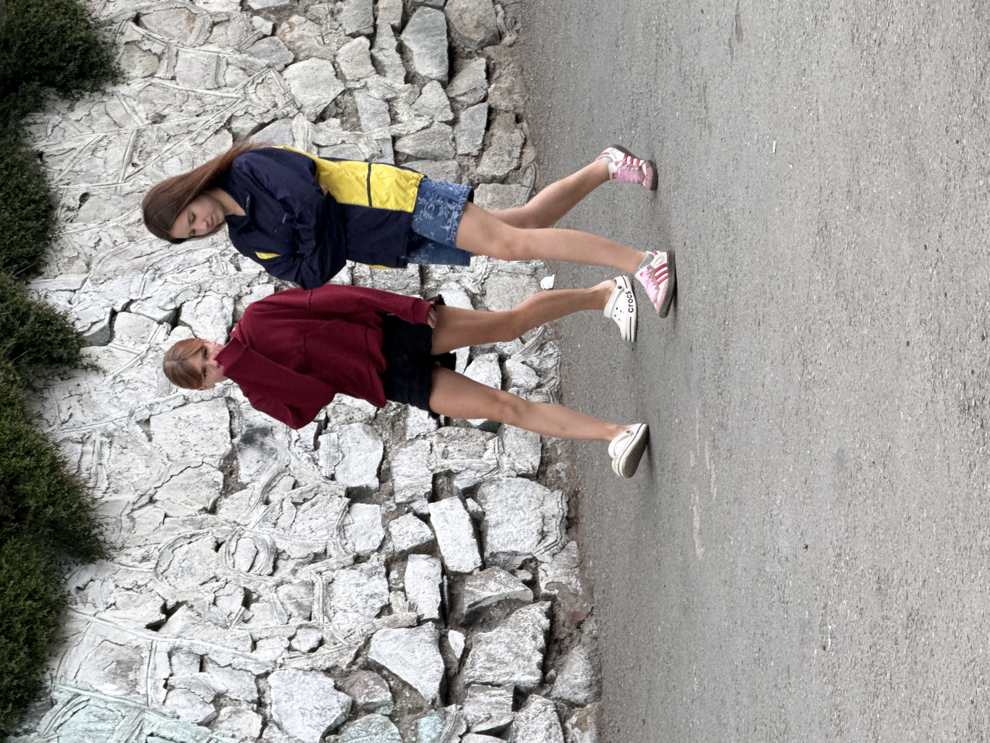 Two young women are standing on a paved surface next to a stone wall. The woman on the left is wearing a dark blue jacket with yellow accents, blue denim shorts, and pink and white sneakers. She has long, brown hair and is looking down. The woman on the right is wearing a maroon hoodie, black shorts, and white Crocs. She has short, blonde hair and is looking forward. Both women appear to be teenagers. The scene is outdoors, and the lighting suggests it could be daytime. There is greenery on the left side of the image. No text is visible.