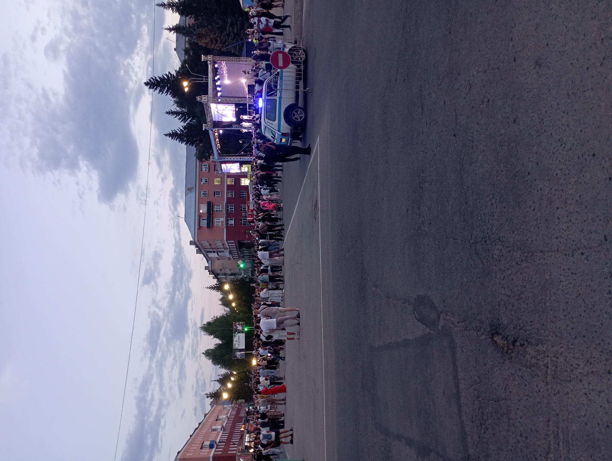 This image depicts an outdoor public gathering in the city of Ridder, Kazakhstan, likely during an evening event. A significant crowd of people, dressed in casual attire, are congregated in a paved open space, possibly a town square or plaza.
In the background, a stage setup is visible with bright lights and screens, indicating a performance or event is taking place. Several buildings, appearing to be multi-story residential or commercial structures, line the sides of the area. Trees are also present, contributing to the urban park-like setting.
A police vehicle with its blue and red lights illuminated is positioned in the foreground, suggesting a security presence. A police officer stands near the vehicle, facing away from the camera. A red and white prohibitory sign is mounted on a temporary barrier, further indicating crowd control measures.
The sky is overcast with clouds, and the overall lighting suggests it is dusk or early evening. The ground is paved with asphalt, showing some wear and tear. The presence of streetlights and building lights indicates the area is illuminated for the event. The scene captures a moment of public celebration or community engagement in Ridder.