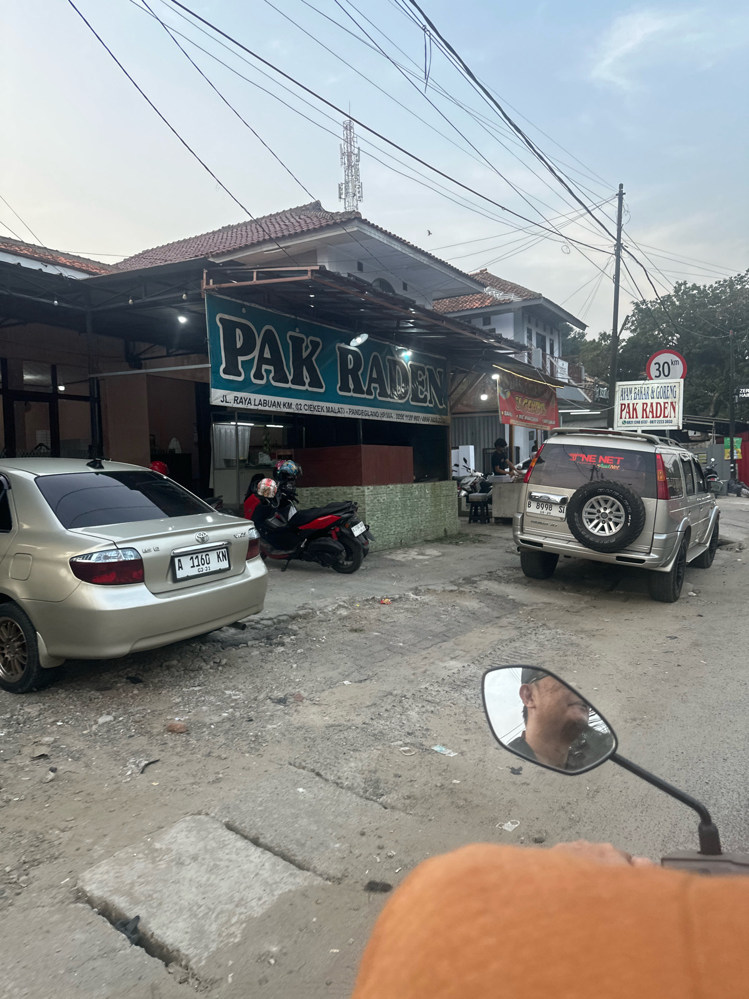The image depicts a street scene in Saruni, Indonesia, with a focus on a roadside eatery named "PAK RADEN". The eatery has a teal sign with white lettering. A silver sedan is parked on the left side of the street, and a red motorcycle is parked in front of the eatery. A silver SUV with a spare tire mounted on the rear is parked on the right side of the street. A speed limit sign indicating "30 km" is visible on a pole. The ground is unpaved and dusty, with some cracks and concrete slabs. In the foreground, a reflection of a person's face is visible in the rearview mirror of a motorcycle, indicating the photo was likely taken from a moving motorcycle. The sky is overcast, suggesting it might be late afternoon or early morning. The buildings in the background appear to be simple structures with tiled roofs. Several power lines crisscross the sky.