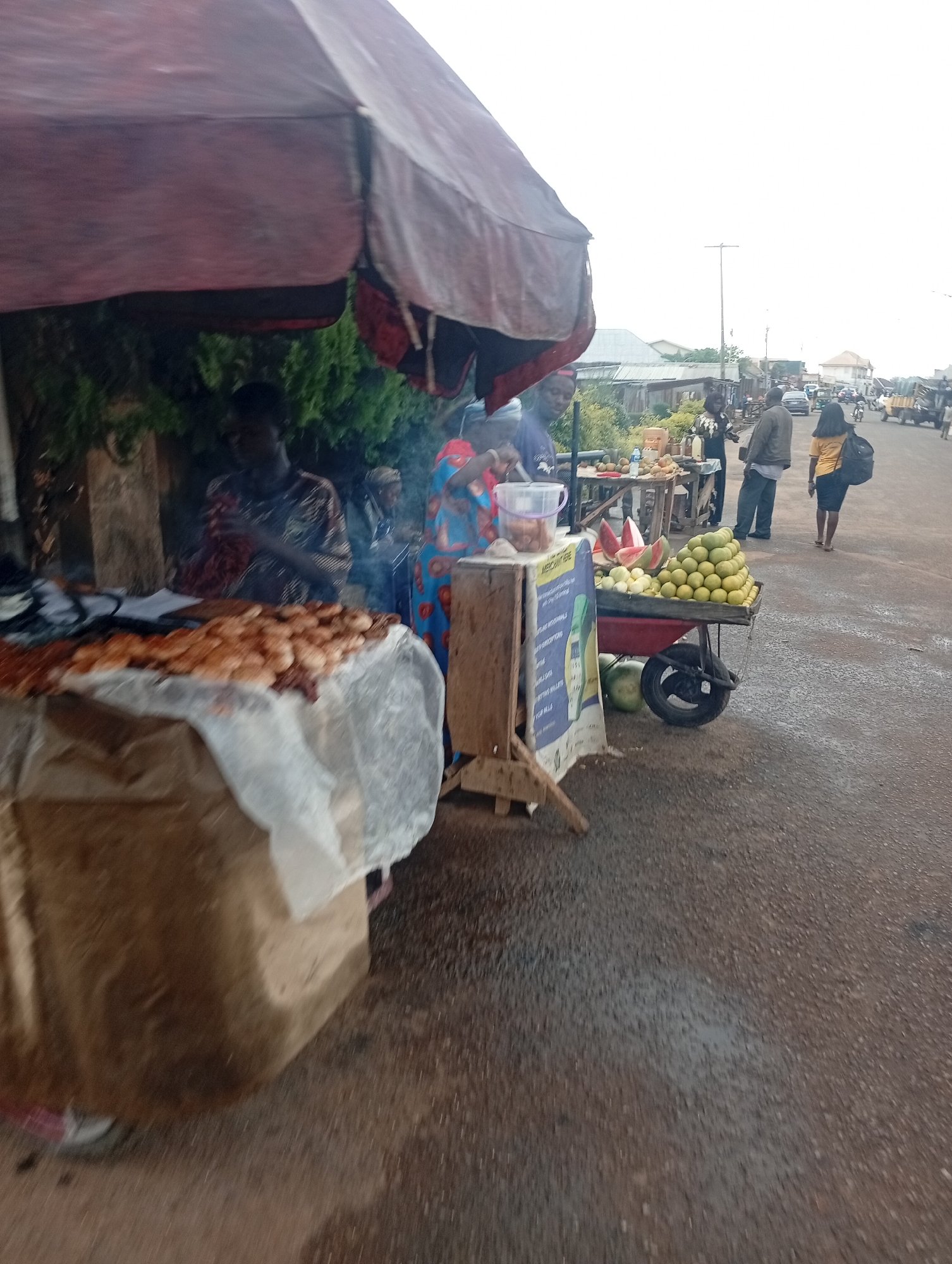 This image captures a vibrant street market scene in Jos, Nigeria, during daylight hours, likely under an overcast sky. The foreground features two distinct vendor stalls set up along a paved road.
On the left, a large, reddish-brown market umbrella provides shade over a food preparation area. Wisps of smoke rise from a table covered with a translucent sheet, displaying what appears to be numerous pieces of grilled or fried food, possibly meat or bread. A person is seen tending to this stall, partly obscured by the smoke.
Adjacent to this, on the right, is a two-wheeled pushcart laden with a large mound of green, round fruits (possibly oranges or grapefruits) and several cut slices of watermelon. A whole watermelon rests on the ground next to the cart. A blue and white sign, partially legible with text including "MEEC...", is attached to the front of the cart, likely serving as an advertisement or product information. Behind the cart, another wooden stall holds more fruits, and a woman in a black top is attending to it.
Several people are visible on the street. A man in a dark jacket and trousers stands near the fruit stalls, seemingly interacting. Further down the road, a woman in a yellow top carrying a dark backpack walks away from the market area. In the background, the street is busy with other pedestrians and vehicles, including cars and a motorcycle, suggesting a lively urban or semi-urban setting. Simple buildings line the distant street. The road surface appears to be asphalt, and the overall atmosphere reflects a typical daily market activity.