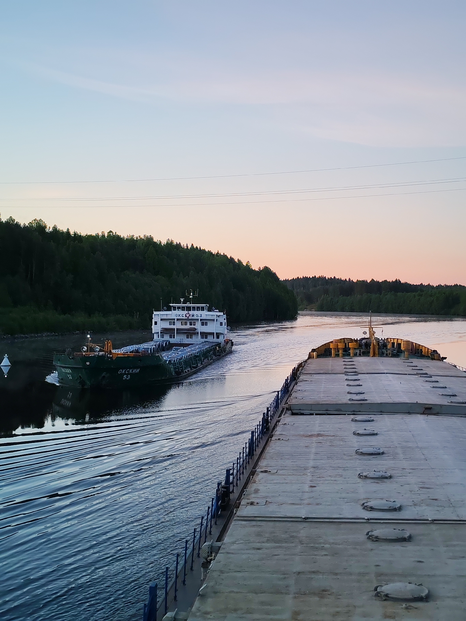 The image captures a peaceful scene on a waterway, likely a river or canal, at either dawn or dusk. The sky displays a gentle gradient from pale blue at the top to soft pink and orange closer to the horizon, indicating early morning or late evening light.
In the mid-ground, a dark green cargo vessel with a prominent white superstructure is navigating the calm waters, creating a visible wake. The name "ОКСКИЙ" (Okskiy) and the number "53" are clearly inscribed on its hull in white. The superstructure also bears the text "ОКСКИЙ И-53". This vessel appears to be a river barge. A white conical buoy is visible to the left of the passing ship.
The photograph is taken from the perspective of another large vessel, the deck of which dominates the foreground on the right side of the image. This deck is flat, grey, and features a series of circular, sealed hatches, bordered by a blue railing. This vessel also appears to be a cargo barge.
Both sides of the waterway are lined with dense, dark green forest, suggesting a natural or canalized environment cutting through a forested area. Overhead power lines are faintly visible against the sky. The water is smooth, reflecting the surrounding landscape and sky. The visible Russian text on the ship, "ОКСКИЙ", points to a location within Russia, such as Depo. The scene evokes a sense of tranquil passage through a serene natural setting.