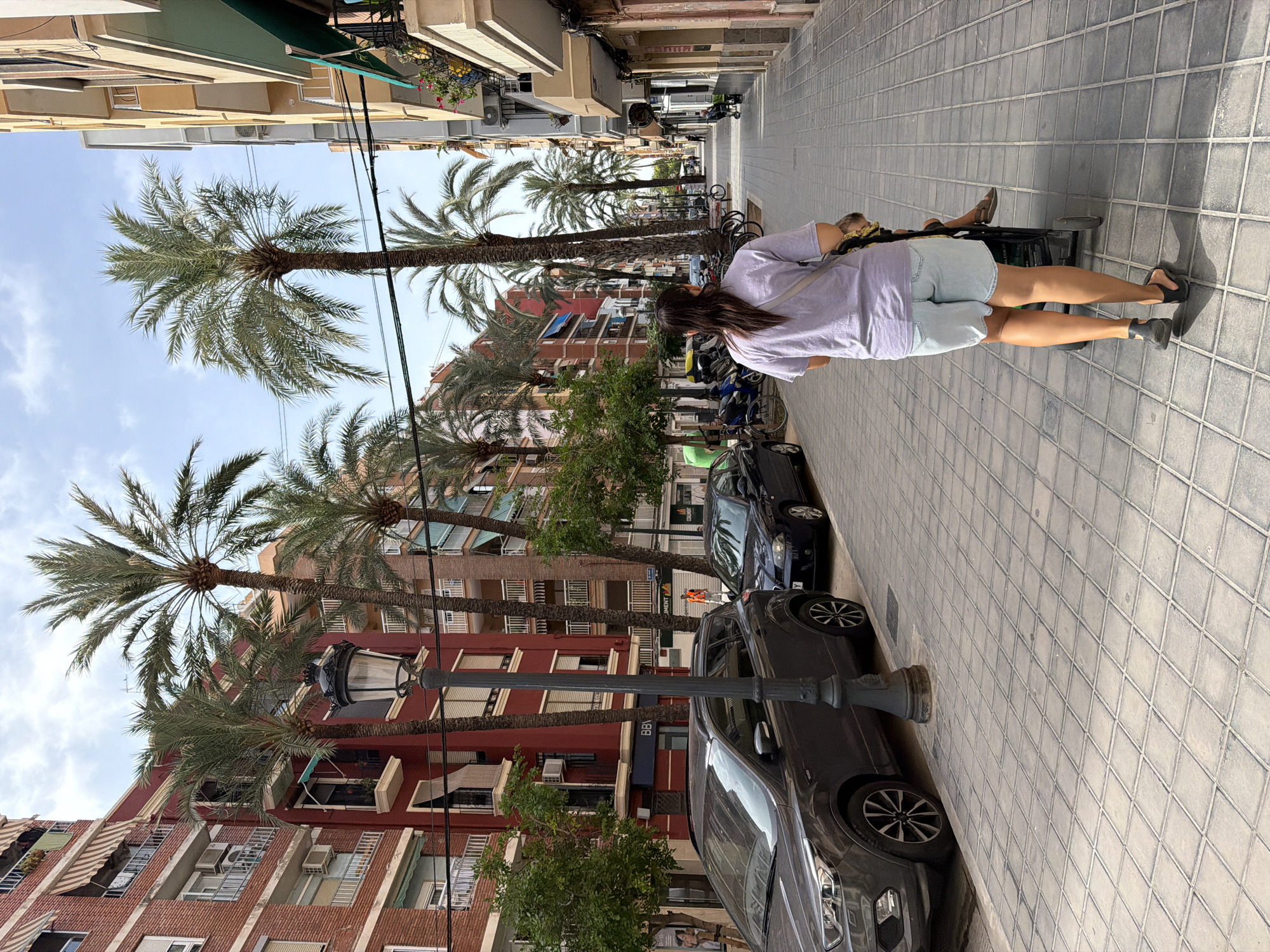 The image depicts a street scene in an urban environment. In the foreground, a woman with long dark hair is walking away from the camera, pushing a stroller. She is wearing a light purple t-shirt and light blue denim shorts, with black sandals on her feet. A strap from a bag is visible across her shoulder. In the stroller, a child wearing sandals is visible. The woman and stroller are walking on a tiled sidewalk.
To the left of the sidewalk, there are parked cars, including a dark gray SUV in the immediate foreground and a dark blue car further down the street. Behind the cars, there are multi-story brick buildings with balconies and awnings.
To the right of the sidewalk, there are shops and businesses with awnings. Further down the street, more buildings are visible, and palm trees are planted along the sidewalk, casting shadows on the ground. The sky is partly cloudy. The overall impression is of a sunny day in a European-style city.