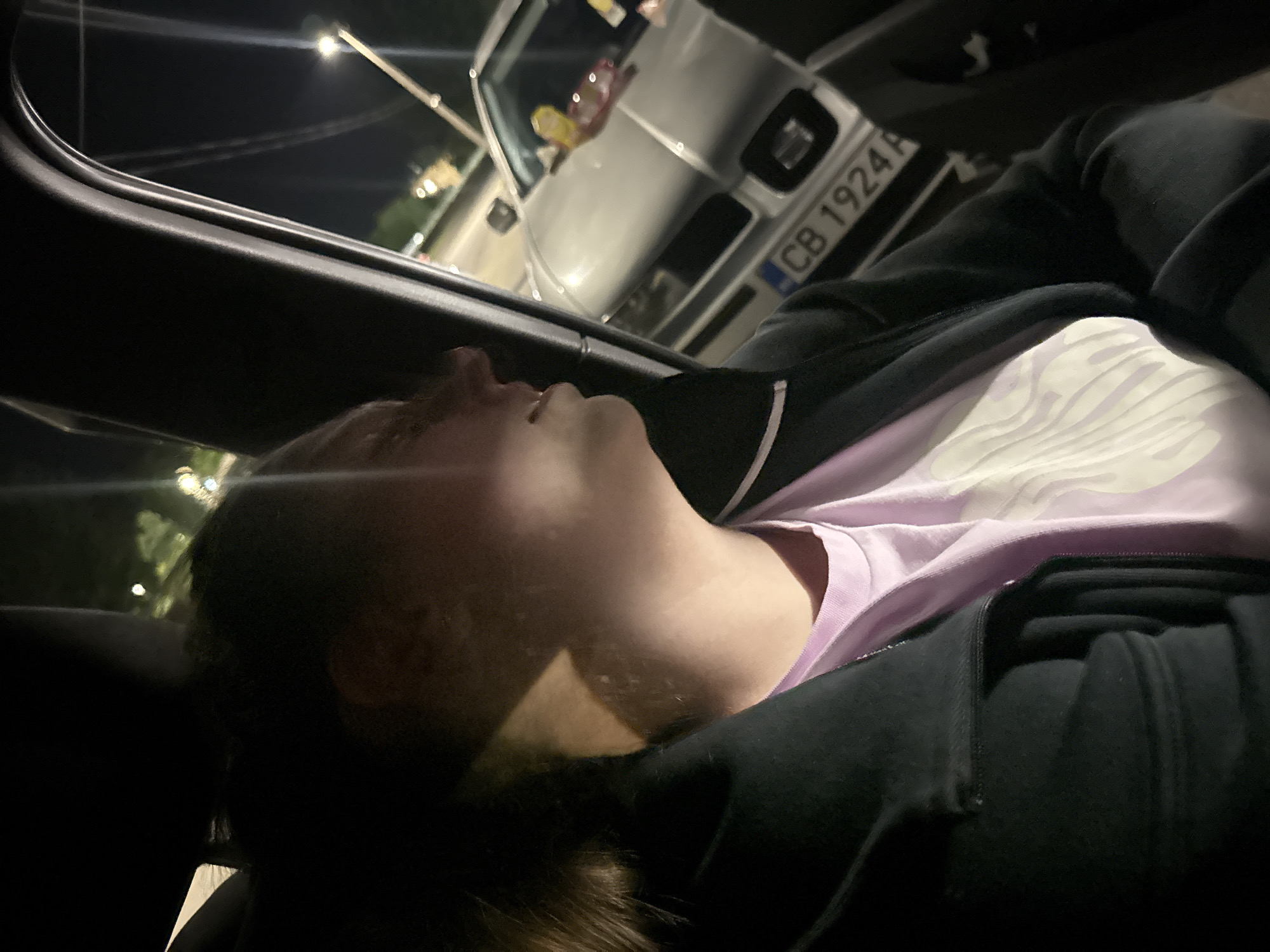 The image captures a nighttime scene observed from inside a vehicle, looking out onto a city street in Kyustendil, Bulgaria. In the left foreground, a young adult, likely female, is visible in profile, facing towards the right side of the frame. Her features are partially illuminated by an unseen light source, highlighting her cheek and neck, with her mouth slightly agape. She has dark hair and is dressed in a dark, possibly black, jacket or hoodie worn over a light purple t-shirt that displays a subtle white pattern on its front.
Outside the vehicle's window, the environment is dark, punctuated by the glow of streetlights in the background. A silver or light grey passenger car is visible in the mid-ground, positioned at an angle, displaying its front portion. The vehicle's license plate is clearly readable as "CB 1924 P," indicating its registration in Sofia, Bulgaria. Through the front windshield of this vehicle, several colorful items, possibly confectionery or decorations, are present on the dashboard or passenger side. The surrounding area suggests an urban setting with some indistinct foliage in the distance.