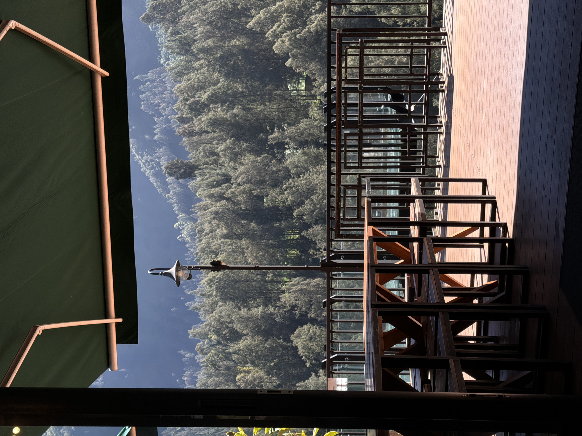 The image depicts an outdoor seating area with wooden tables and chairs on a wooden deck, overlooking a dense forest and a mountain range. A dark green awning extends over the seating area, providing shade. A black lamppost with a glass lantern stands near the railing. The railing is made of black metal bars. The forest appears lush and green, with sunlight filtering through the trees. The mountain range in the background is covered in trees and appears to be hazy. The image appears to be taken during daylight hours, with strong shadows cast by the tables and chairs on the deck. The location is identified as Jetak, Indonesia, suggesting a rural or natural setting within the city. There are no people or animals visible in the image. The overall scene conveys a sense of tranquility and natural beauty.