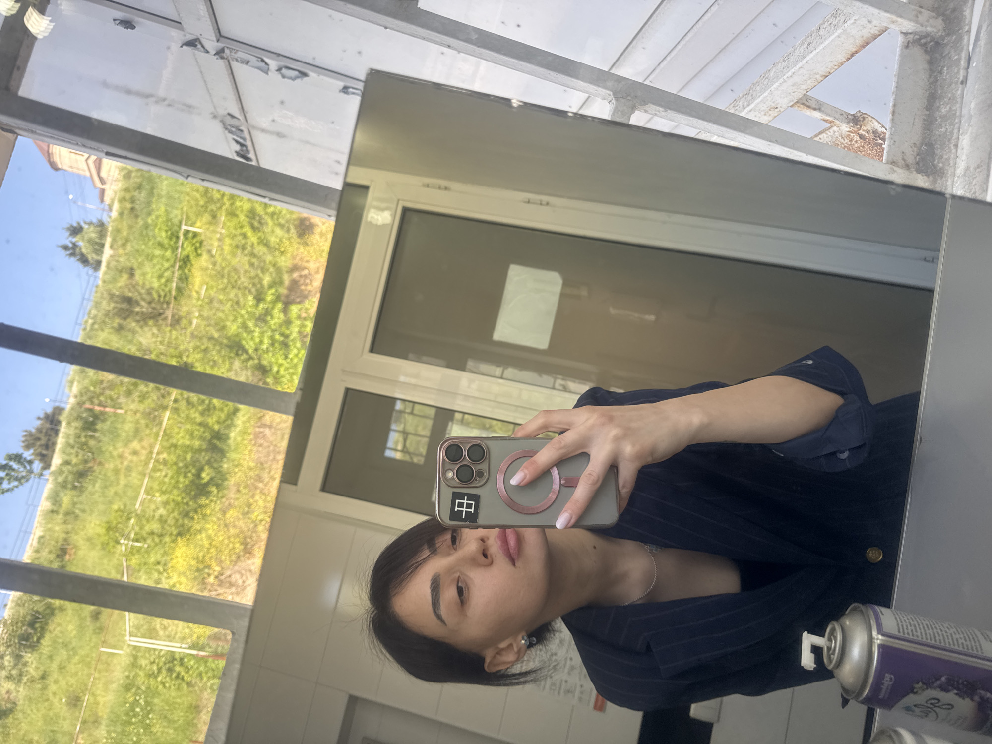 A selfie, taken indoors, features a young woman holding a smartphone, reflecting in a mirror. The phone, a gray iPhone with three camera lenses, has a magnetic ring on the back and a sticker with a Chinese character on its left side. The woman is wearing a navy blue pinstripe blazer over a dark top, with a silver necklace visible.
In the lower right corner of the mirror, two aerosol cans are partially visible. The reflection in the mirror reveals a window showing an exterior view of a grassy area with trees and a blue sky, suggesting a park or natural landscape. The mirror's surroundings show white wall tiles and metal framework, suggesting a bathroom or restroom setting.
The location may be Tbilisi, Georgia, considering the description of the area, but no definitive elements confirm this detail.