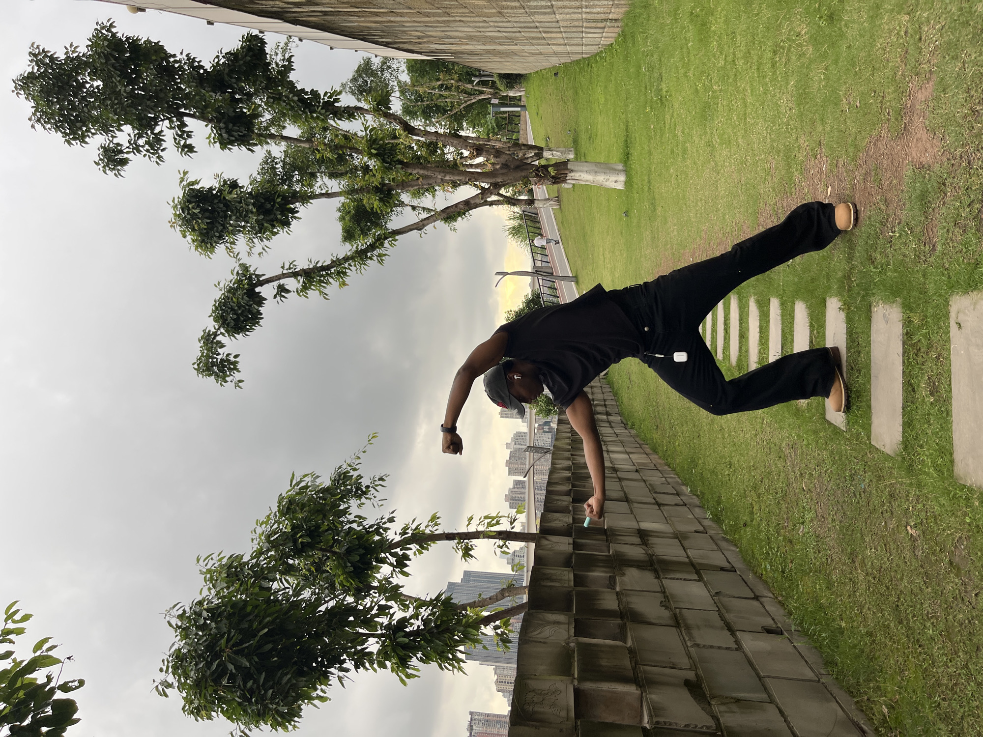A man in black clothes and a baseball cap is posing on a park path in Yuzhong District, China. He is leaning over a low brick wall with his right arm extended, holding a small object, while his left arm is bent with a clenched fist. His legs are extended in the opposite direction, with his feet on the grass and a paved path of concrete slabs. In the background, there are trees, a grassy area, and a row of high-rise buildings. The sky is overcast.