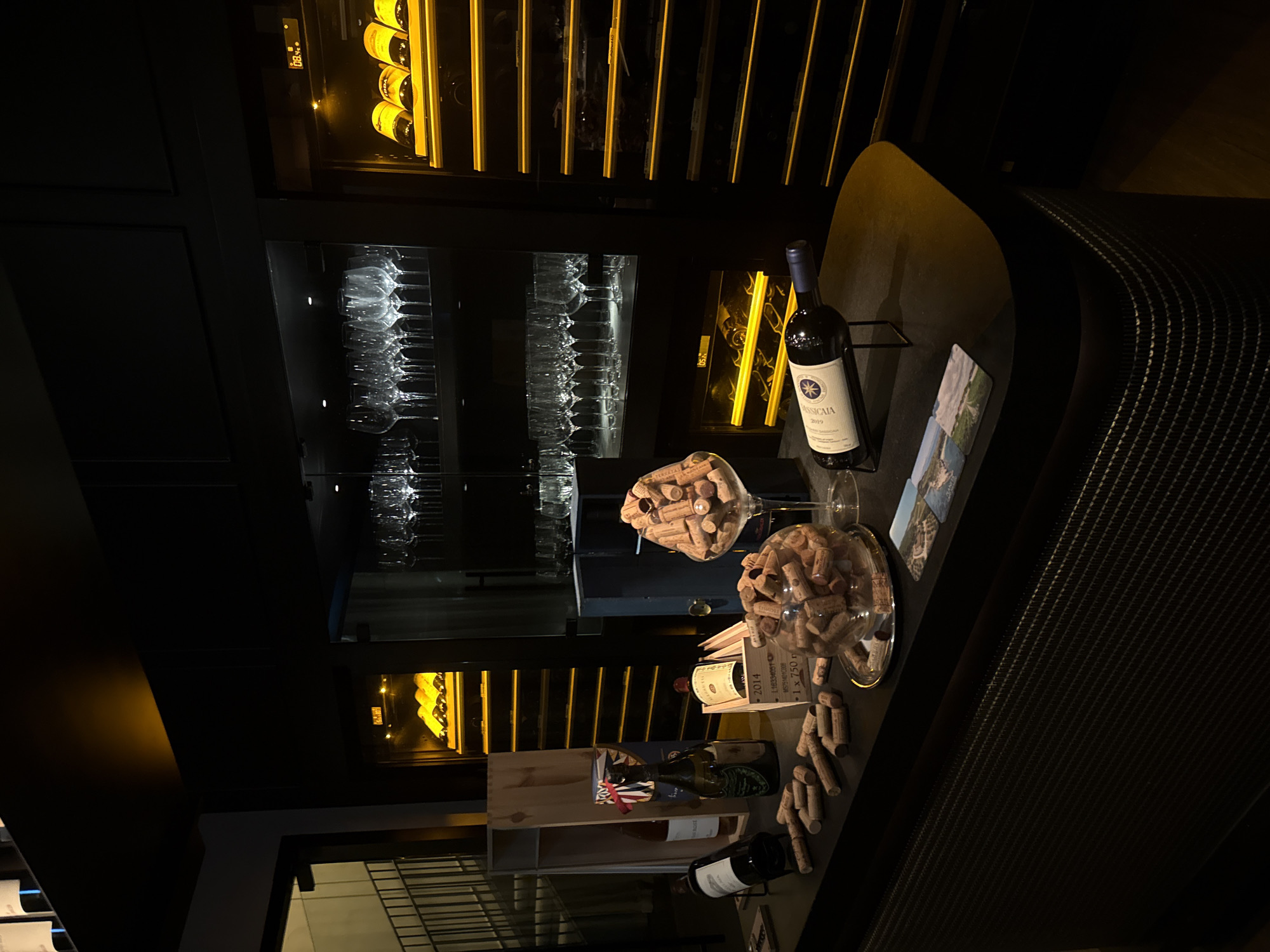The image shows a bar area, possibly in Dubai, United Arab Emirates. A wine refrigerator containing bottles of wine and glassware is in the background. A bottle of Sassicaia 2019 wine and two glasses filled with corks are on a counter next to the refrigerator. A wine rack with bottles of wine and related items is below the counter. The scene is dimly lit.