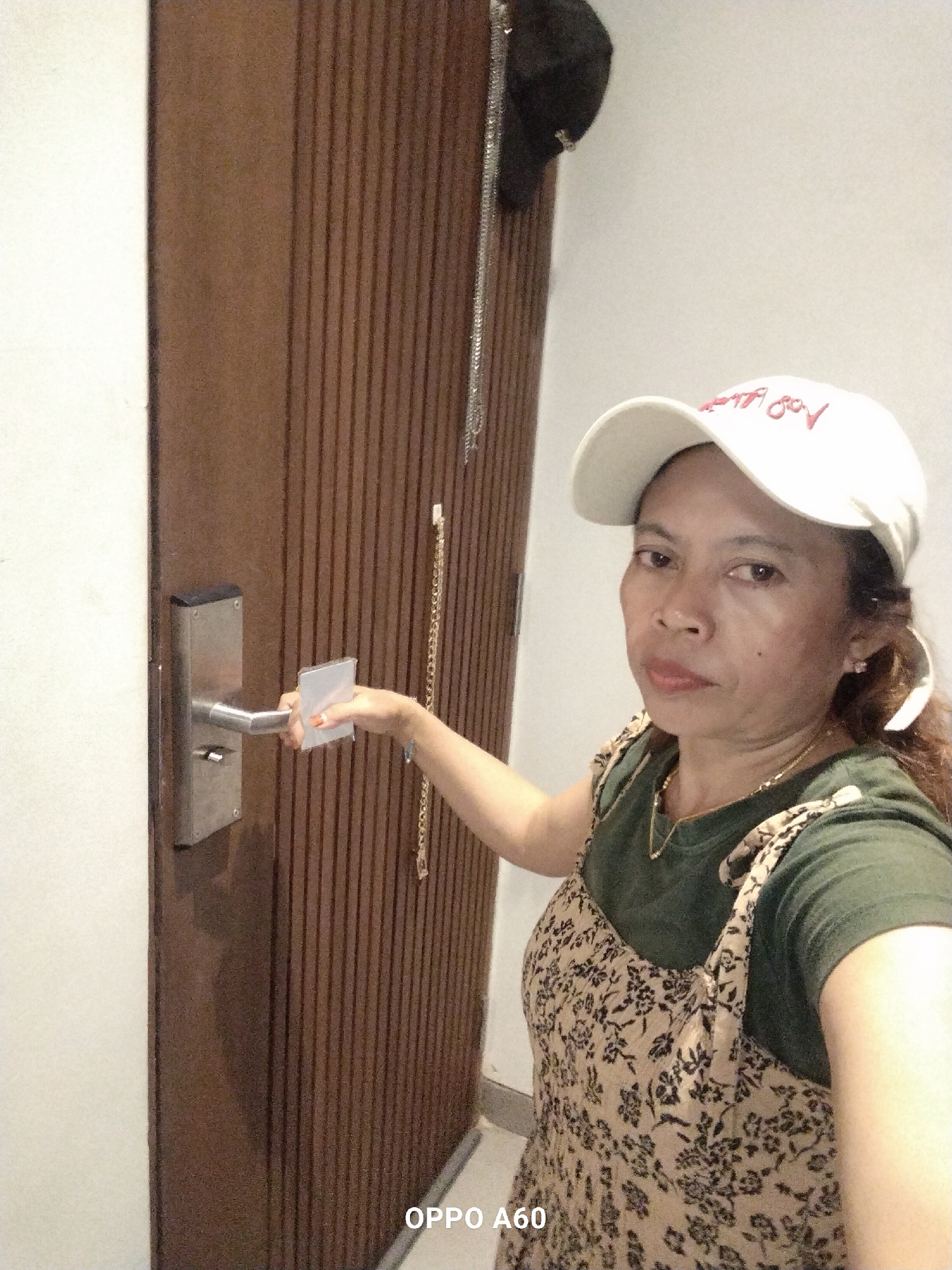 An image taken by a smartphone shows a woman wearing a white baseball cap and a floral-patterned dress over a green t-shirt. She is standing in front of a wooden door with vertical slats. Her right hand is holding a rectangular object and is extended towards the door handle, which is a silver lever. A black baseball cap hangs from a hook on the door. The environment appears to be indoors, likely a hotel room or apartment in North Kuta, Indonesia, indicated by the "OPPO A60" watermark in the lower right corner of the image, which is a model of smartphone commonly used in Indonesia.