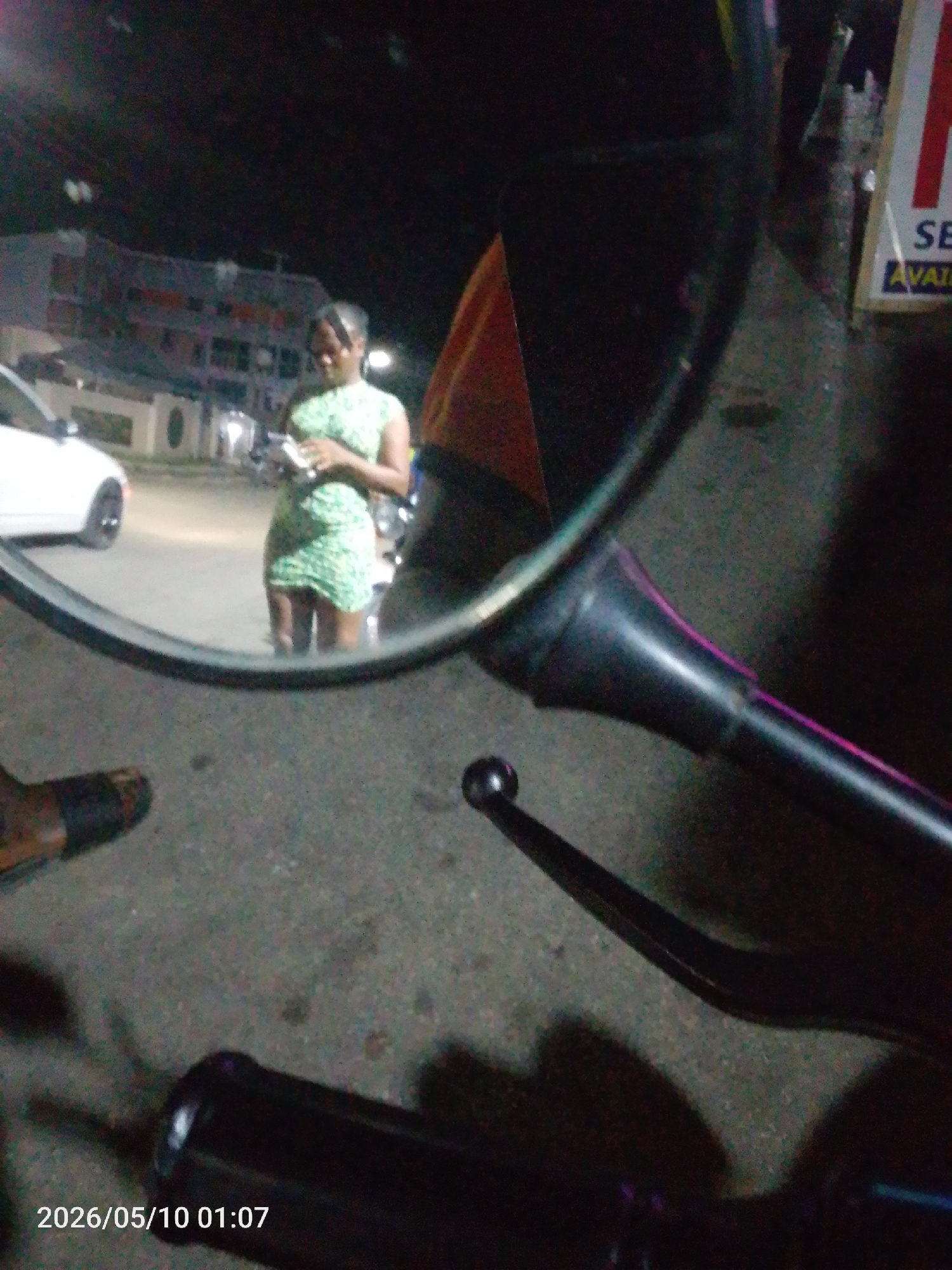 The image appears to be taken from a motorcycle, focusing on the mirror's reflection. The reflection shows a woman in a green dress standing on a street, possibly using a cell phone. A white car and a building are visible in the background. The location appears to be in a city at night, potentially Lokoja, Nigeria, based on the prompt. The immediate foreground includes the motorcycle's handlebars and mirror, along with a foot in a sandal. The overall scene suggests an urban environment at night.