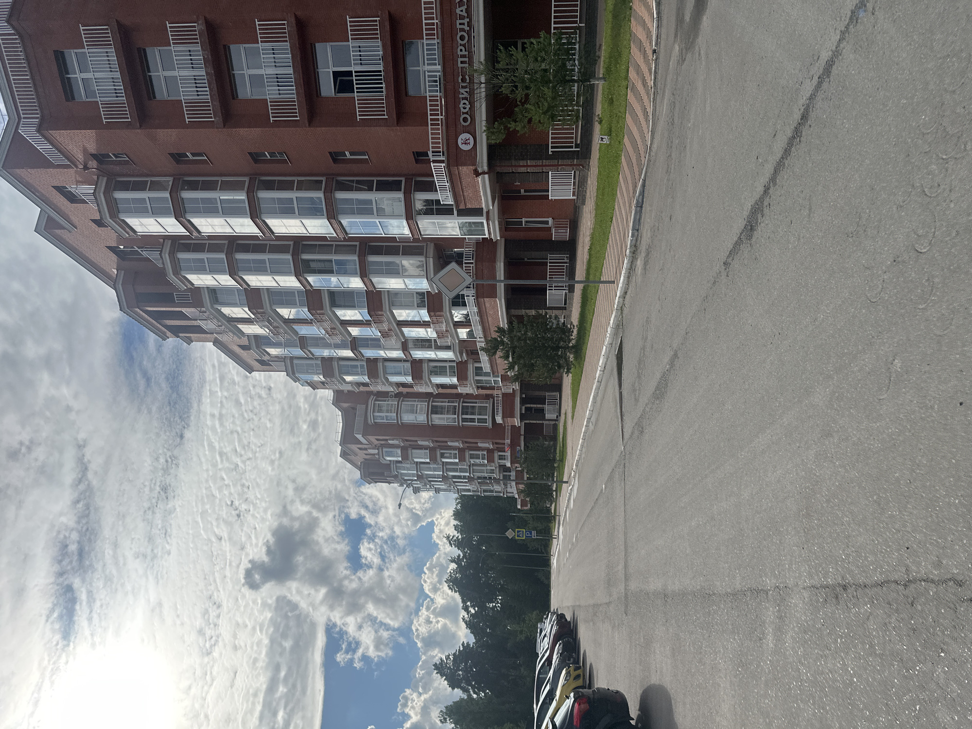The image shows a row of brick apartment buildings with many windows and balconies. In front of the buildings is a paved road with parked cars on the left side. A sidewalk with grass and trees borders the road in front of the buildings. A sign on the building says "ОФИС ПРОДАЖ," which translates to "Sales Office." The sky is cloudy with patches of blue. The location appears to be in Snt Del'fin, Russia, based on the text.