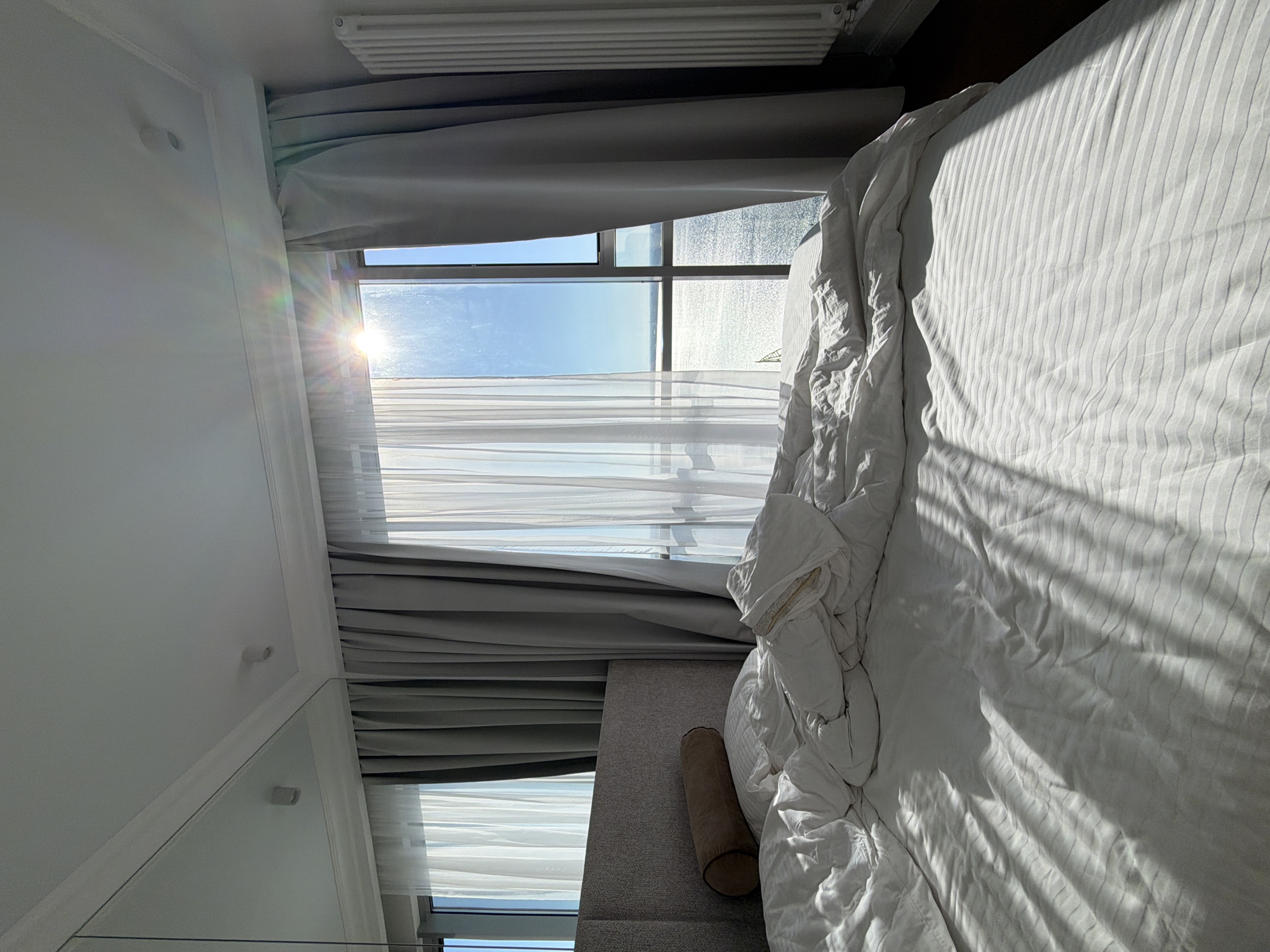 The image shows a rumpled bed in a room with a large window overlooking a body of water. Sunlight streams through the window, creating bright reflections and shadows on the bedsheets. The room appears to be a bedroom, with a headboard visible behind the pillows. The overall impression is one of a recently occupied bed in a bright, airy room with a view. There is no indication of the city of Saratov, Russia, or any specific geographical location.