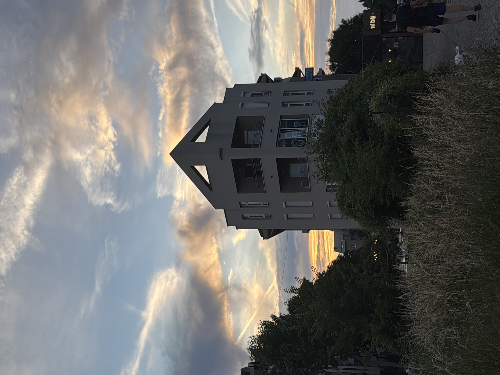 The image depicts an urban twilight scene in Cukarica Urban Municipality, Serbia. The upper two-thirds of the frame features a sky transitioning from light blue to a warm palette of orange, yellow, and pink, illuminated by a setting sun. Scattered clouds are visible, and two distinct airplane contrails form an 'X' pattern in the mid-left sky. In the center-left, a modern, multi-story building with a gray facade stands prominently. It features a distinctive triangular peak at its roofline with an open triangular void below it. The building has multiple windows and balconies. On a lower floor, a window displays graphic images of teeth, indicating a dental practice. To the right of this building, another structure displays a partial sign reading "OMATOLOG," consistent with a "STOMATOLOG" (dentist) sign. Further right, a dark commercial building features illuminated text "EN GMA" (likely "ENIGMA"). The midground contains dense, dark green trees and bushes, particularly on the left. The immediate foreground is filled with dry, tall, light brown grasses. On the right side of the frame, a man in a dark t-shirt and shorts walks away from the viewer, holding a leash. A small, white dog is visible near his feet, slightly ahead of him. The scene is characterized by the warm, golden light of the setting sun, creating contrasts between illuminated clouds and shadowed foreground elements.