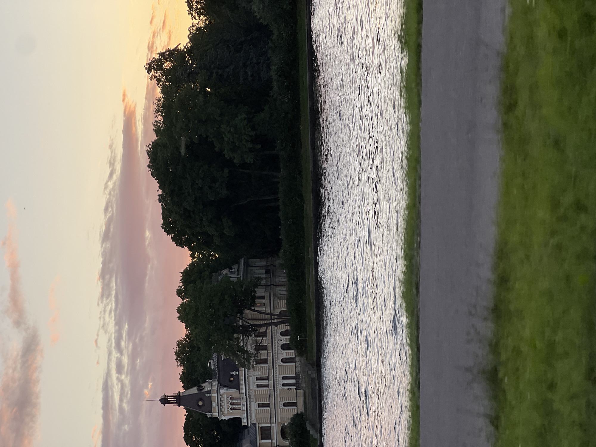 The image depicts a picturesque view of a waterfront along the Neva River in Saint Petersburg, Russia, at dusk. A grand, light-colored, multi-story building with a prominent dark tower stands on the riverbank, partially obscured by lush green trees. The building exhibits architectural details indicative of a historical estate or palace, with arched windows and decorative elements. The Neva River flows in the foreground, its surface rippling and reflecting the soft hues of the sky. The sky above is a canvas of pastel colors, with streaks of pink and orange clouds suggesting the setting sun. The immediate foreground shows a paved path and a strip of green grass, implying a park or promenade area. There are no people visible in the scene.